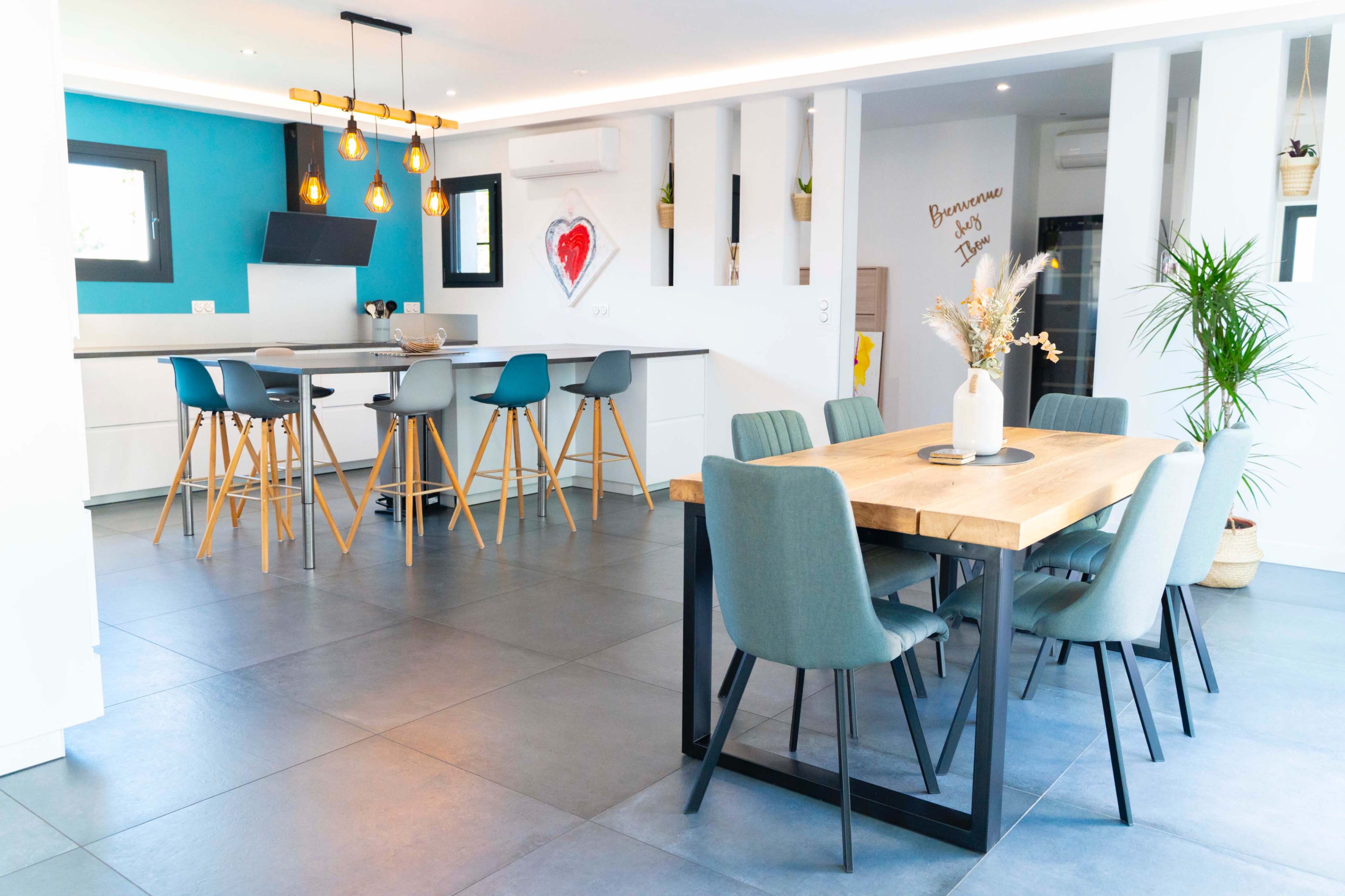 A modern dining area features a wooden table surrounded by six chairs, with a kitchen and living space visible in the background.