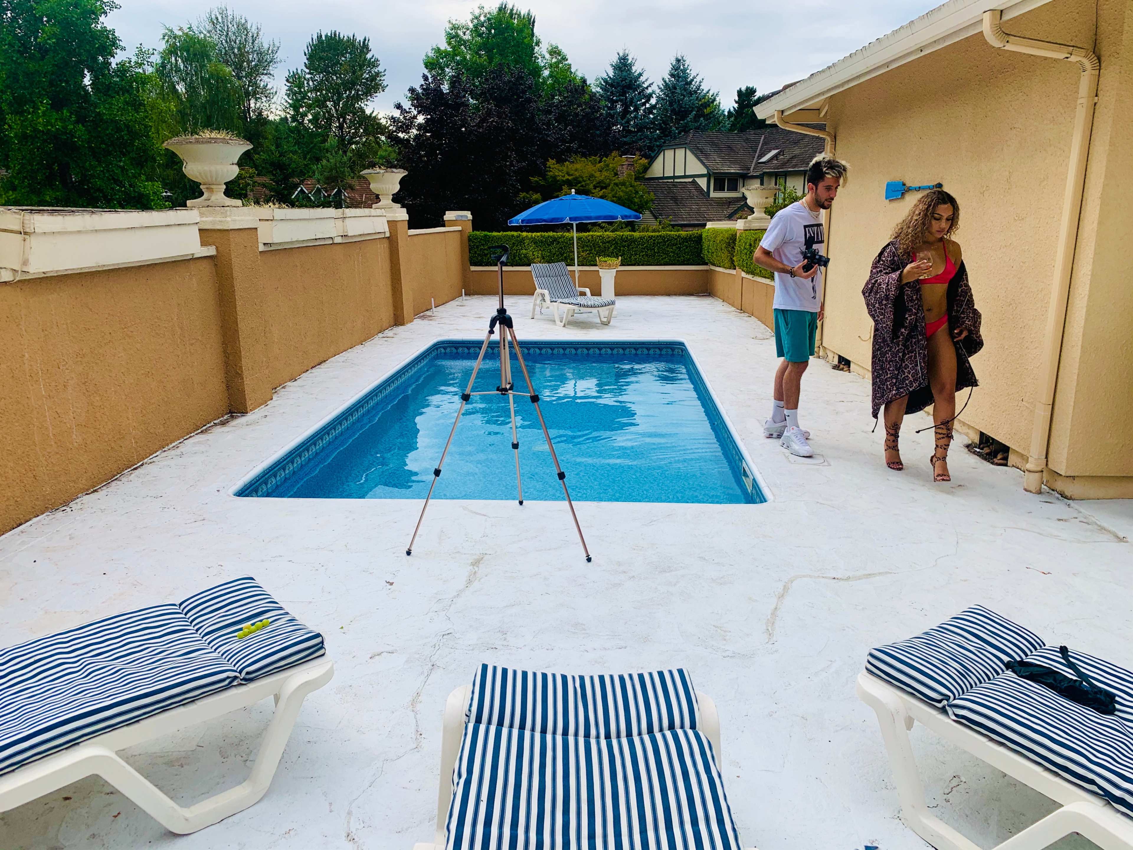 A man and a woman stand near a swimming pool surrounded by lounge chairs and an umbrella.