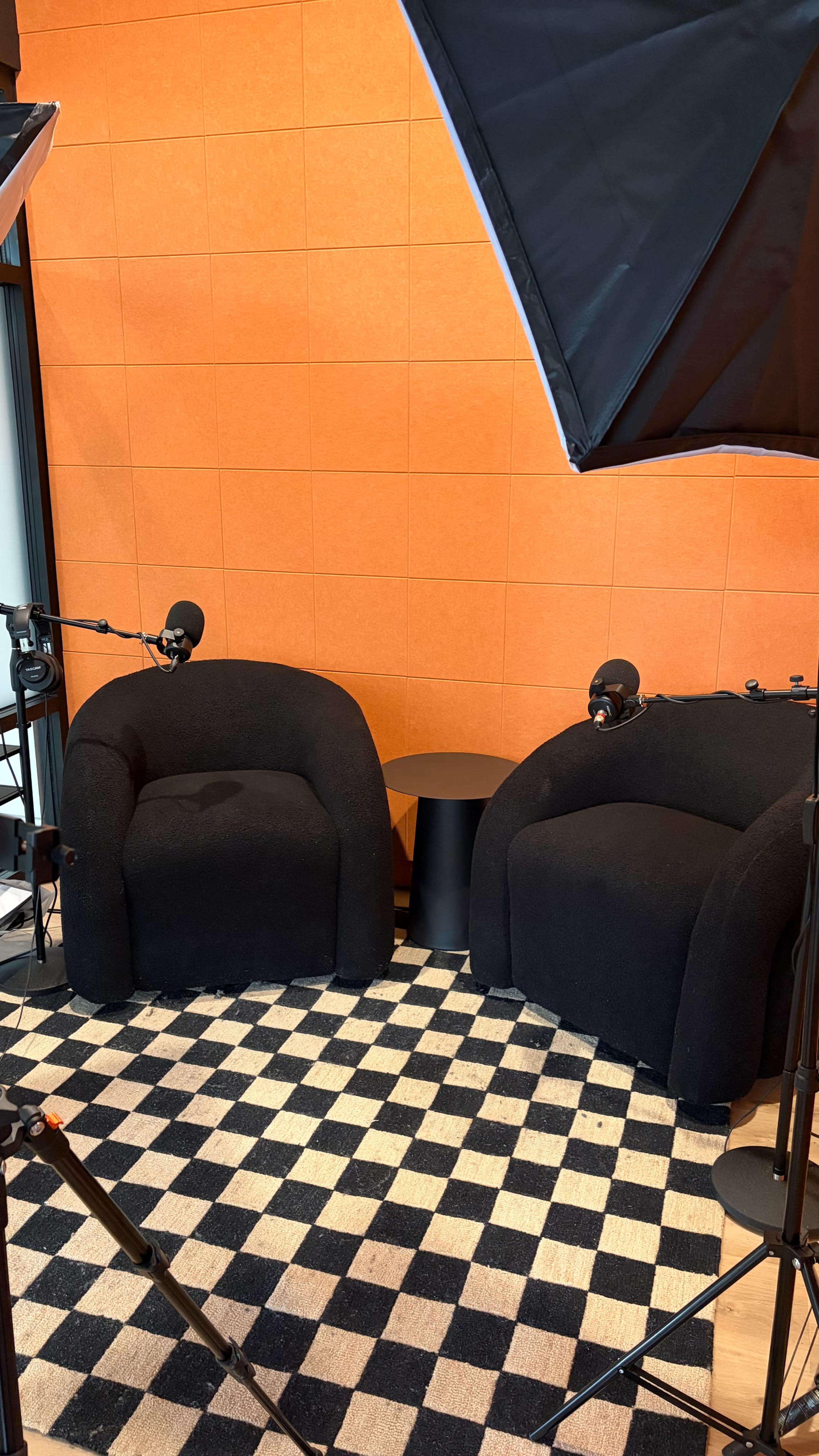 The image shows two black armchairs facing each other on a checkered rug, with an orange wall in the background and microphones positioned nearby.
