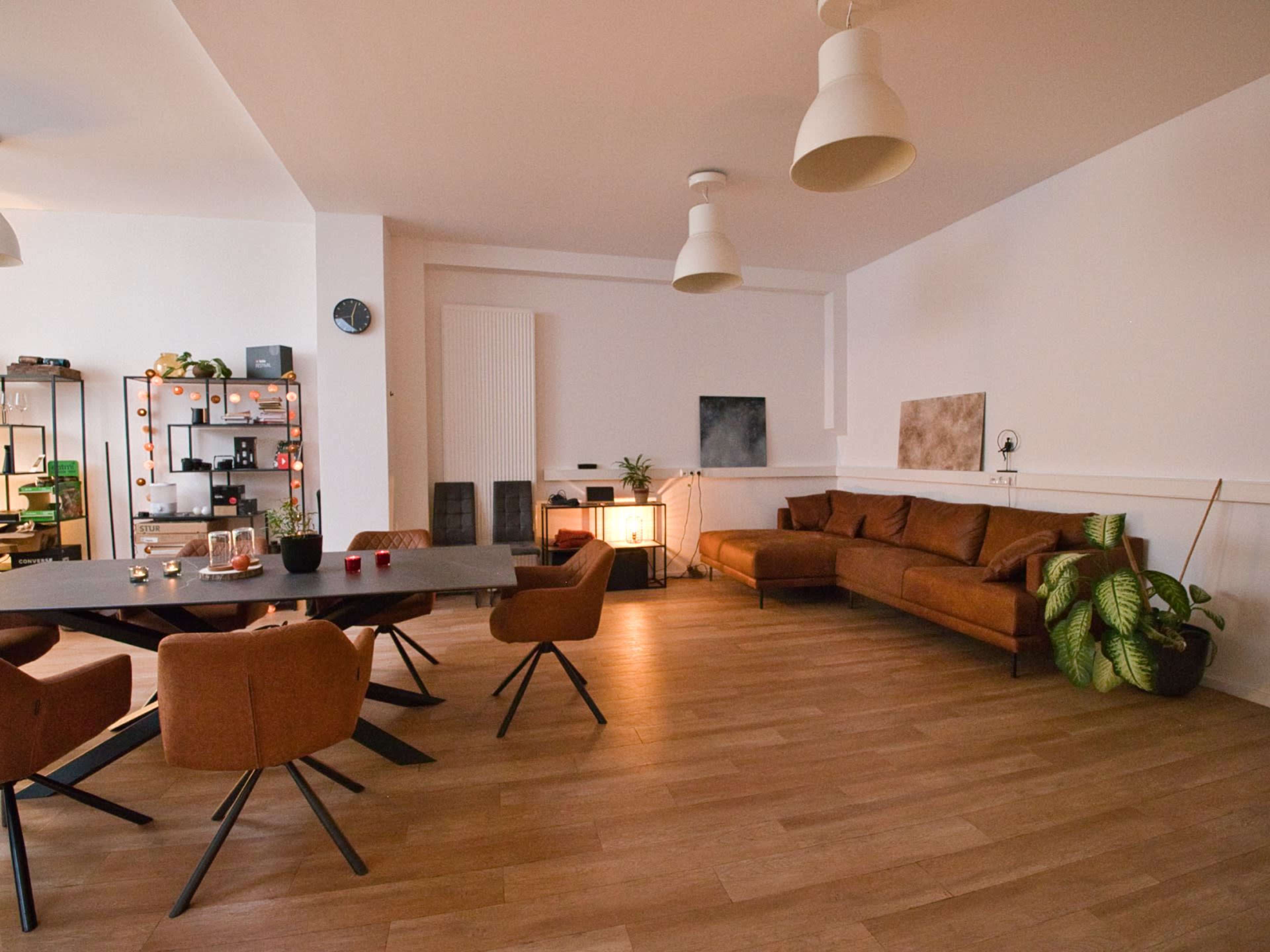 The room features a spacious dining area with a large table surrounded by brown chairs, a sectional sofa, and various plants placed throughout.