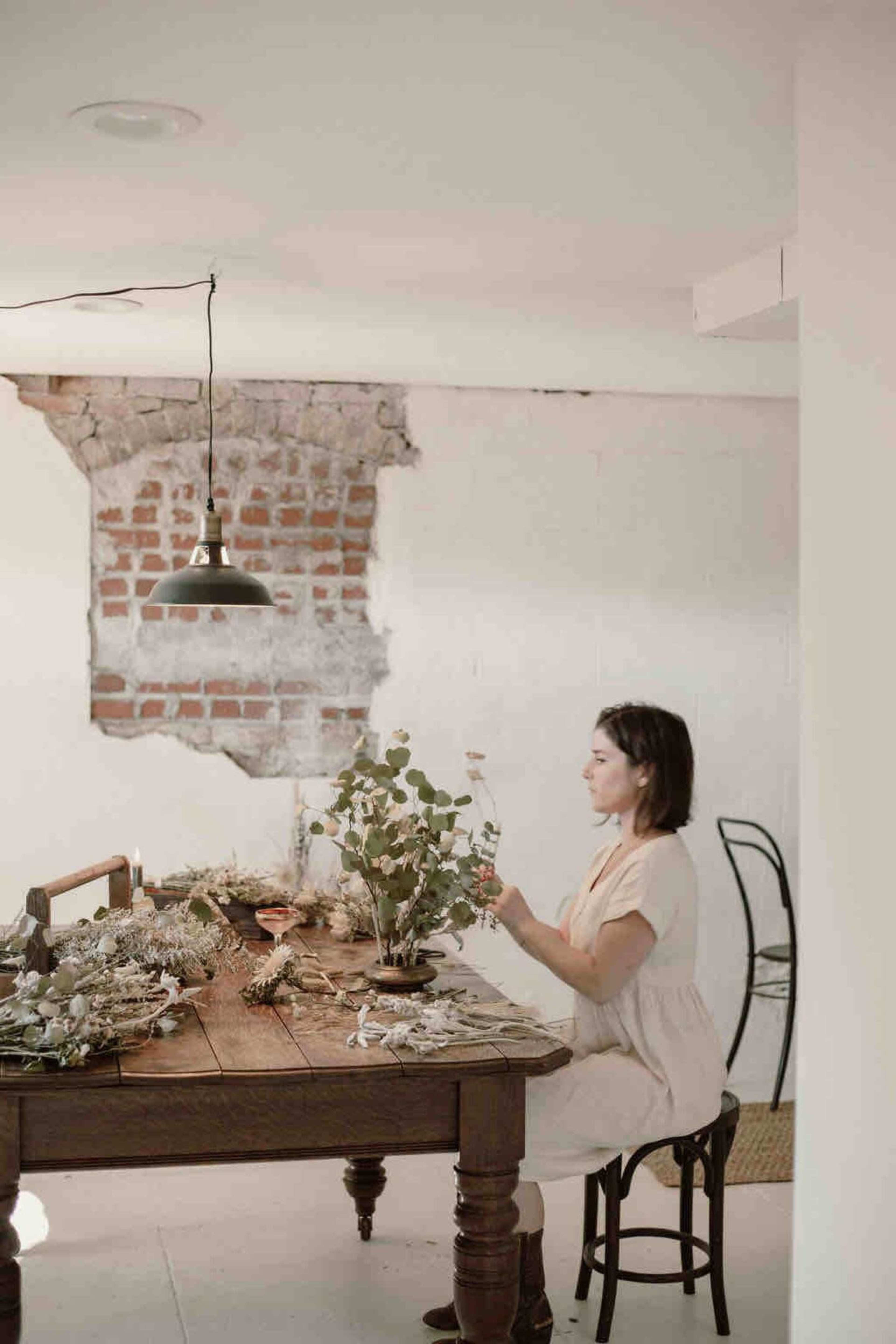 A woman sits at a wooden table adorned with dried flowers, while a brick wall serves as the backdrop.