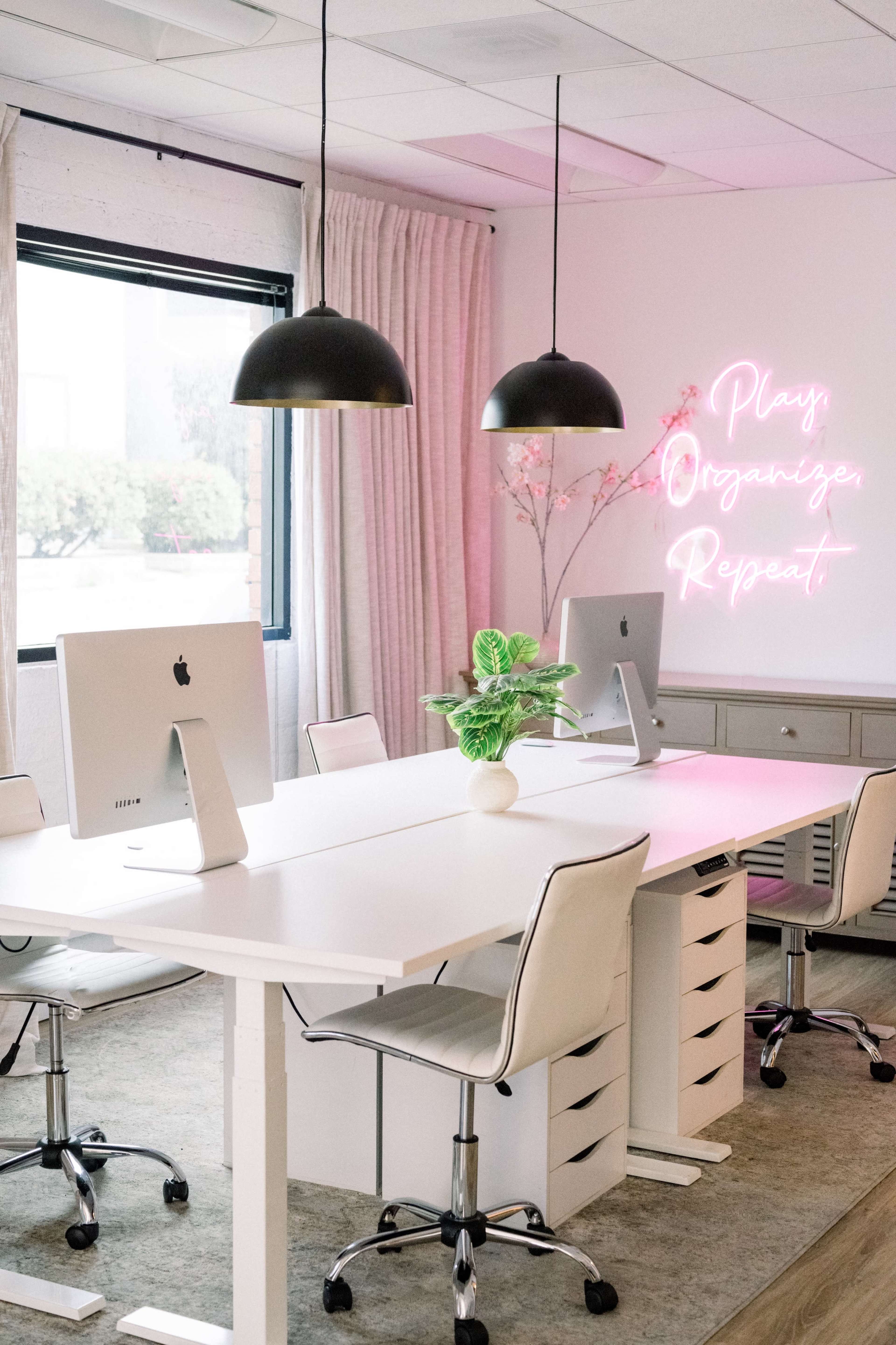The image shows a modern office space with two computers on white desks, a plant centerpiece, and a neon sign that reads "Play Organizer Repeat."