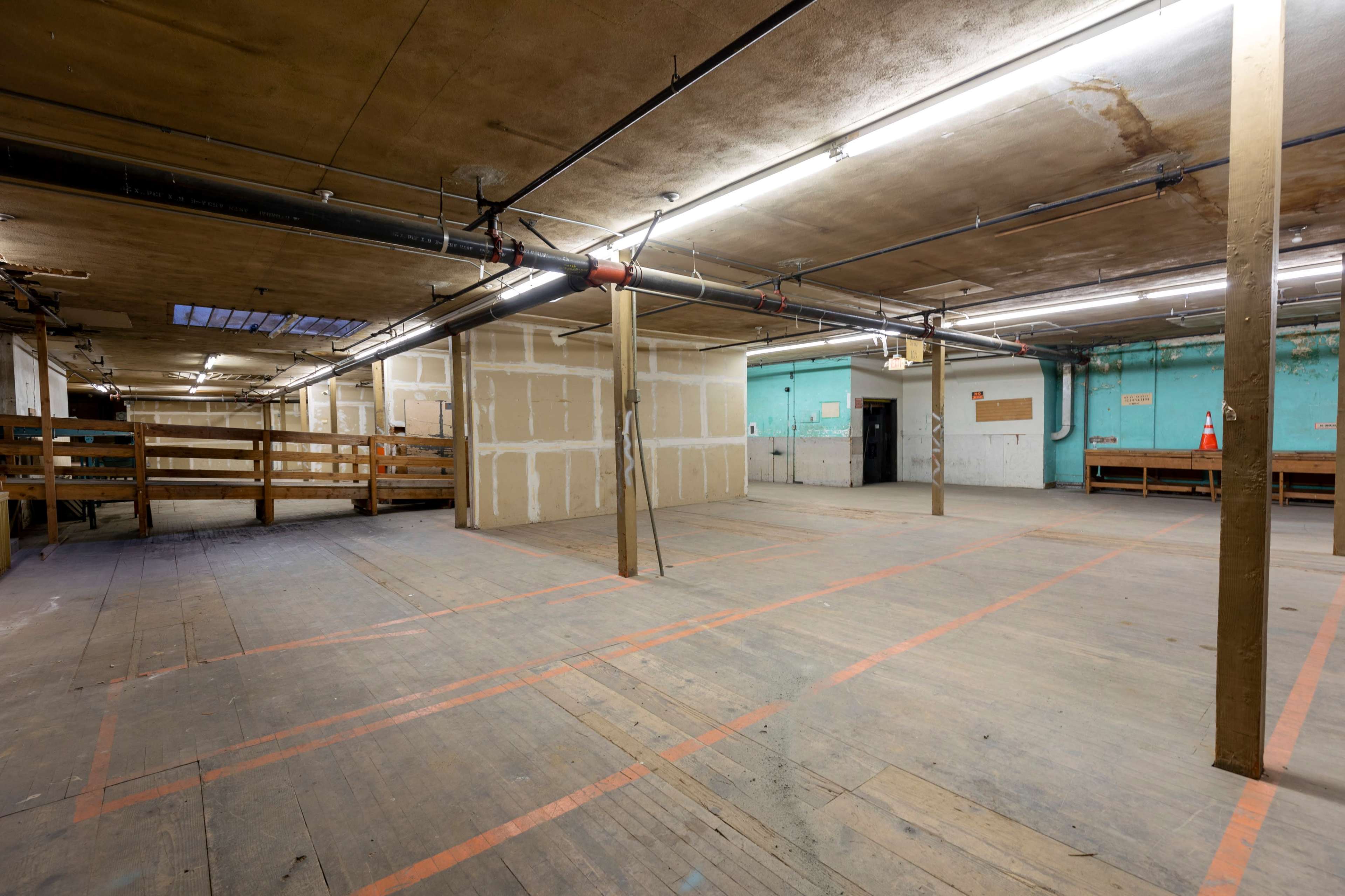 The image shows an empty, dimly lit warehouse interior with wooden floors and bare walls partially covered in beige paint.