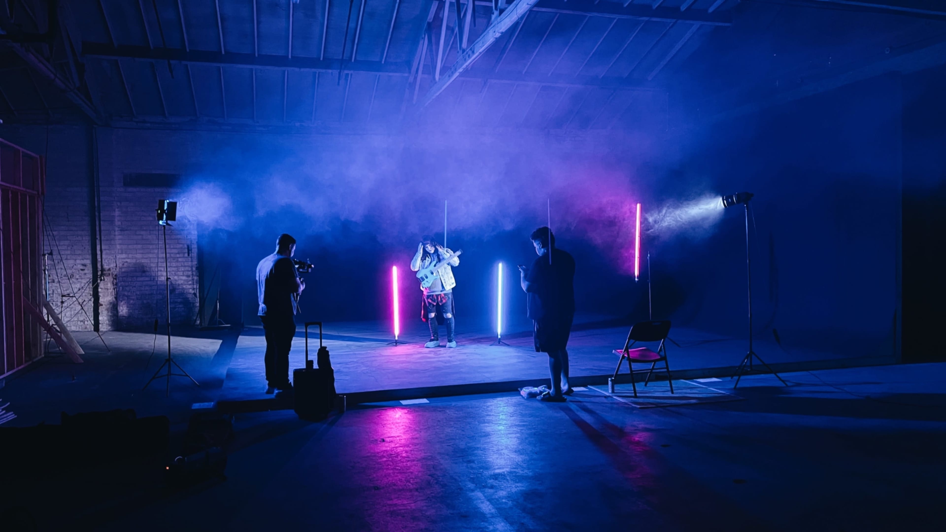 A performer stands on a small stage illuminated by colored lights, while two camera operators capture the scene in a fog-filled warehouse.