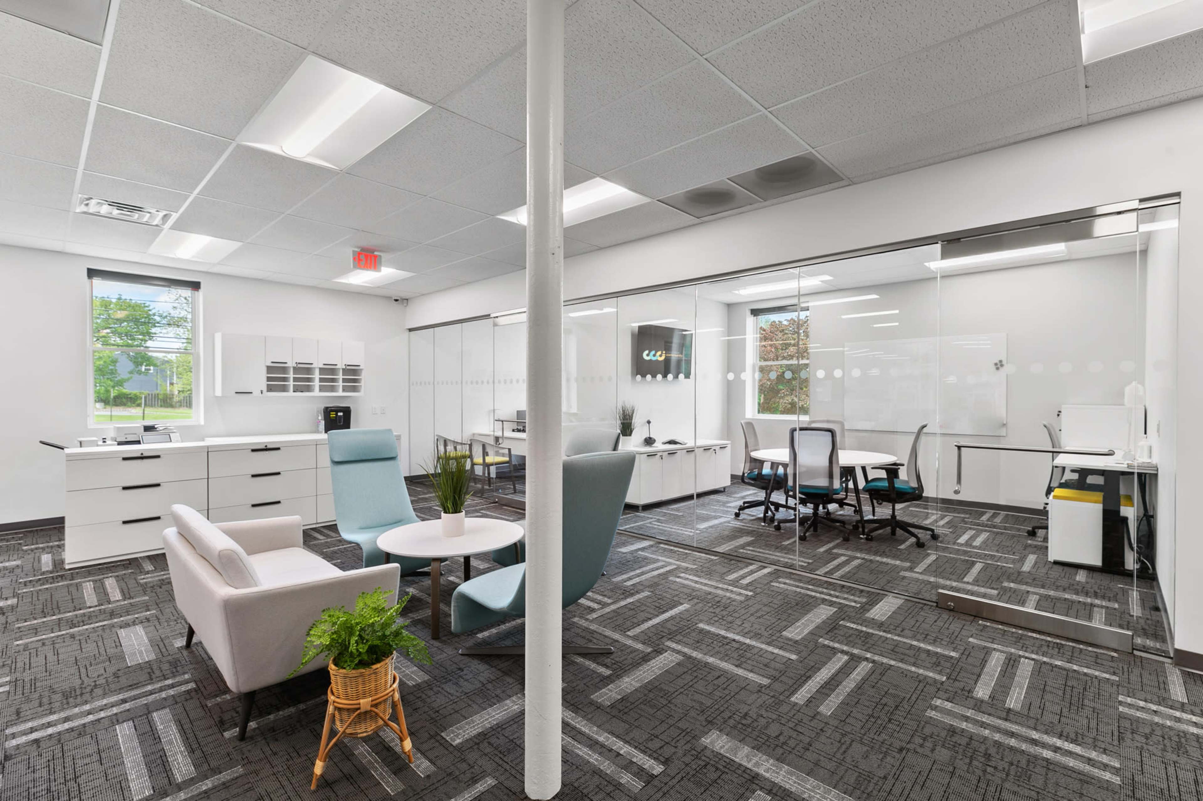 A modern office space featuring a glass wall, a central seating area with a couch and chairs, and multiple workstations with sleek furniture.