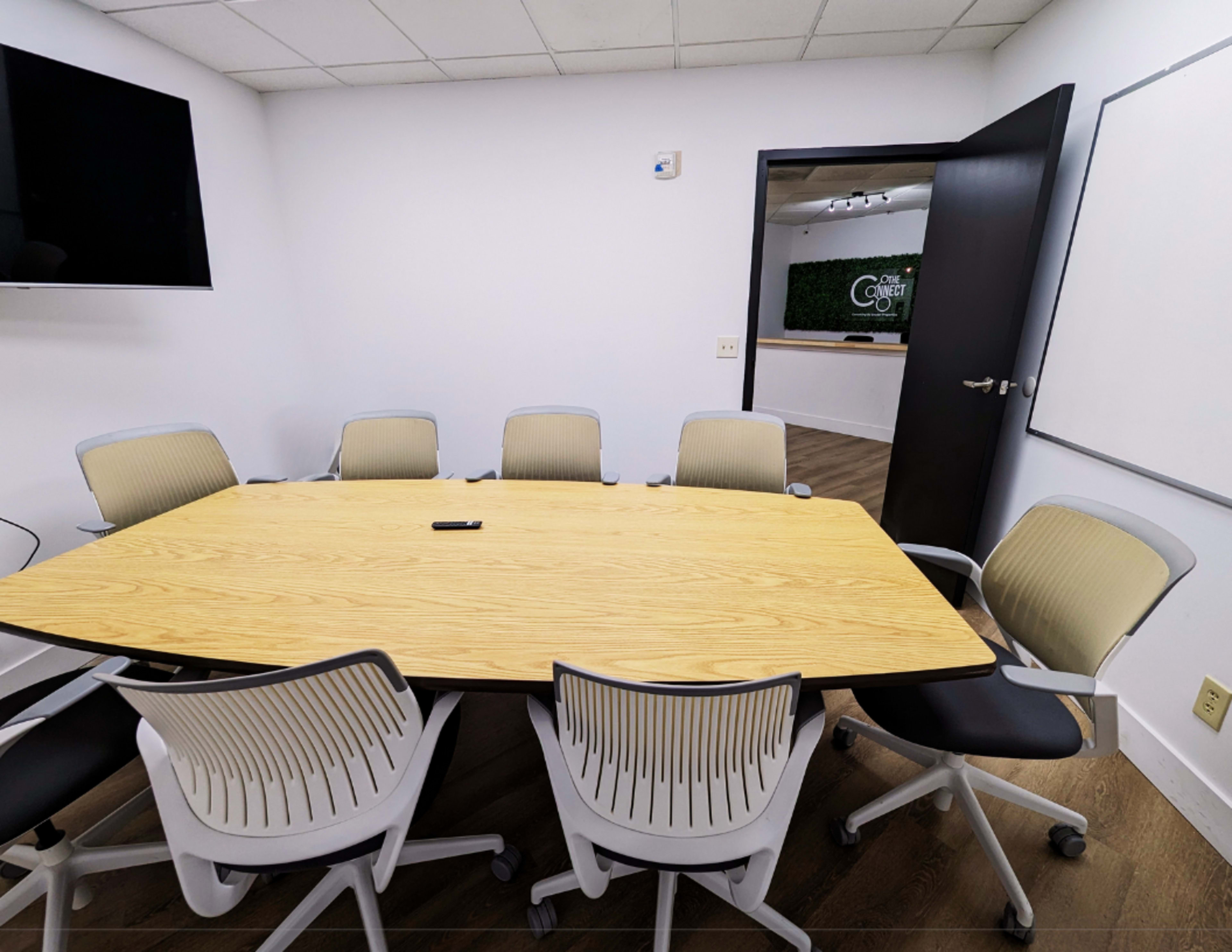 A large, oval wooden conference table surrounded by white and gray office chairs is set in a minimalist meeting room.