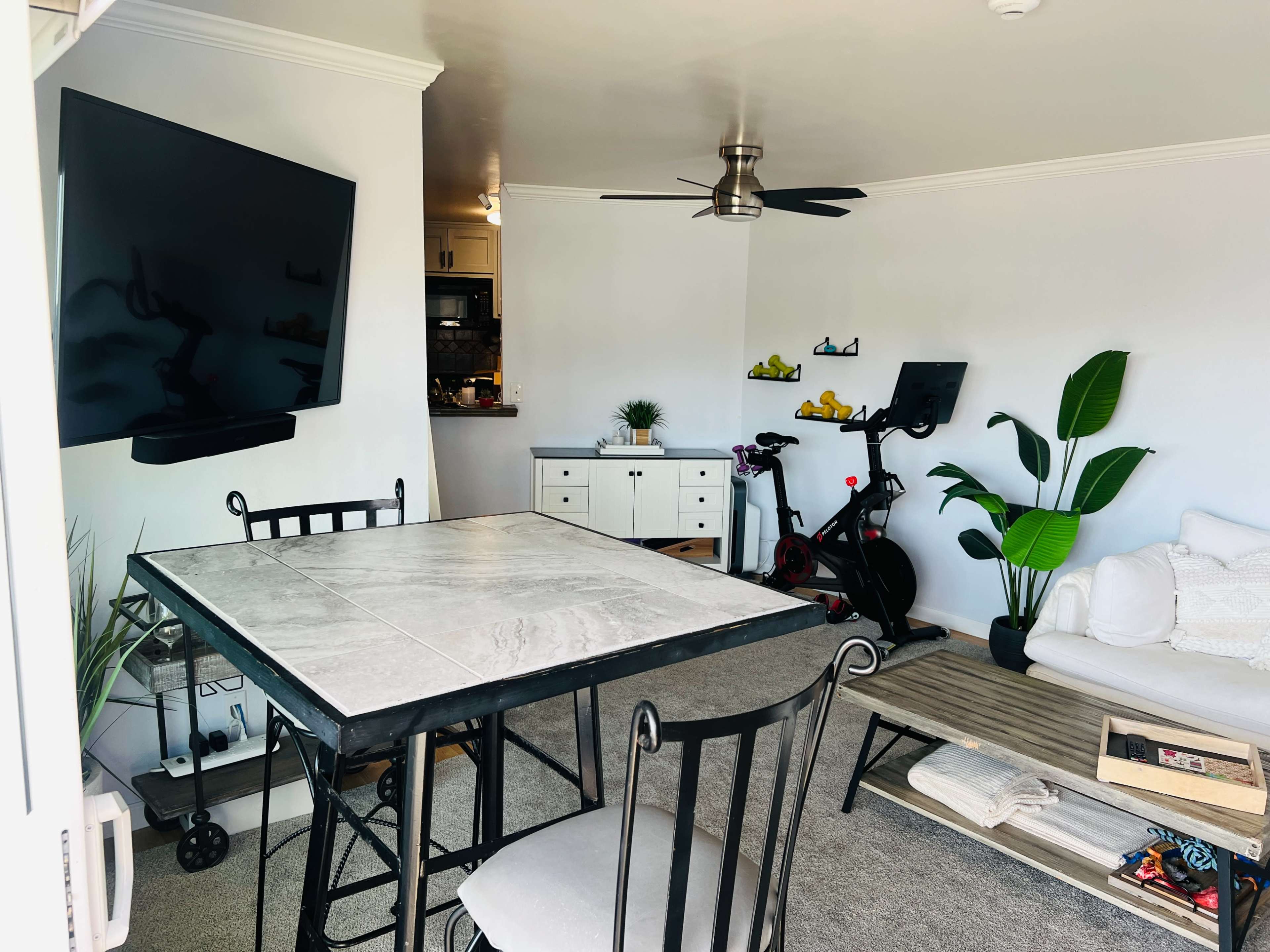 The image shows a modern dining area with a marble table, a large wall-mounted TV, a stationary bike, and a small couch beside a potted plant.