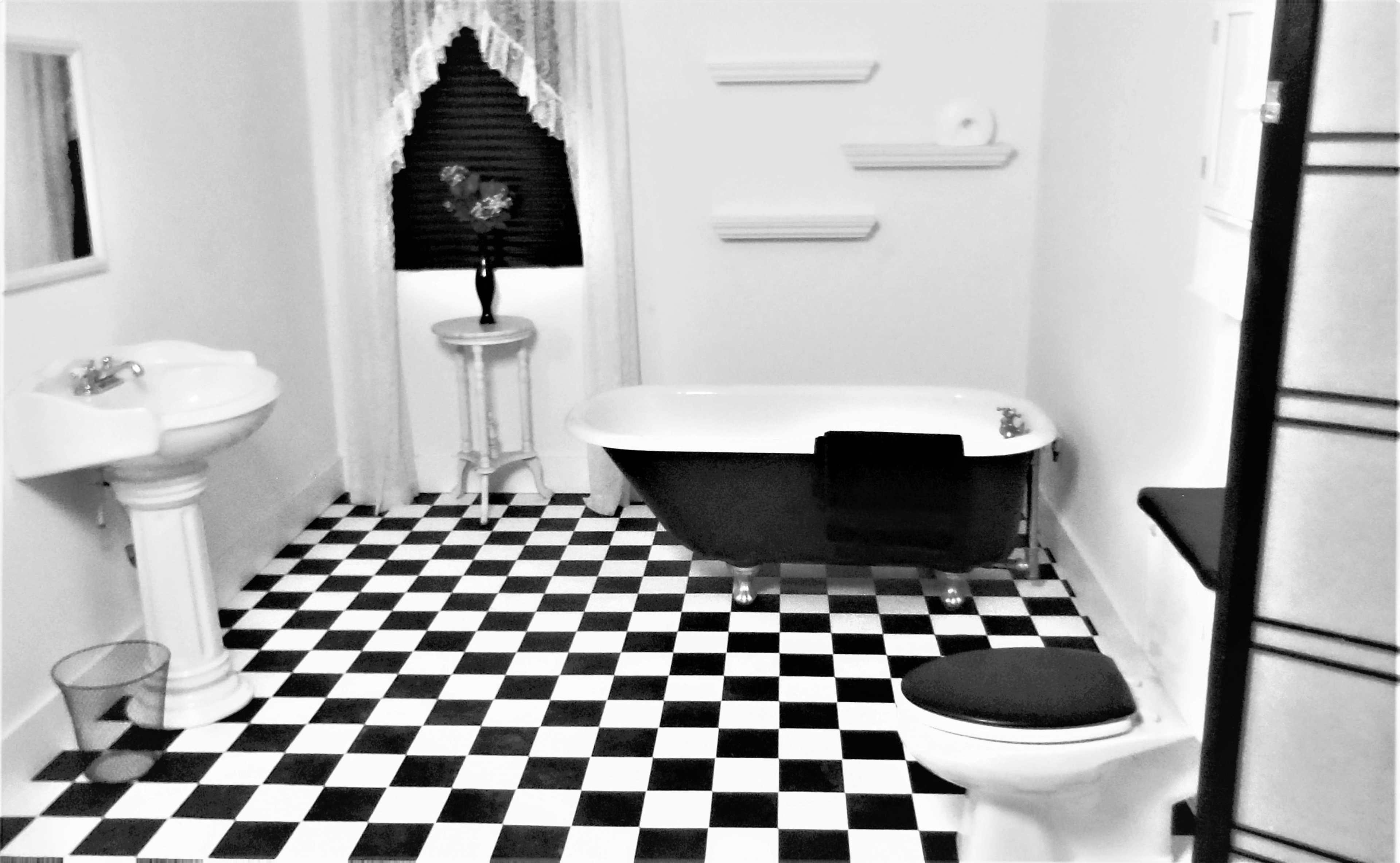 A black and white tiled bathroom featuring a clawfoot bathtub, a pedestal sink, a toilet, and a small decorative table beside the window.