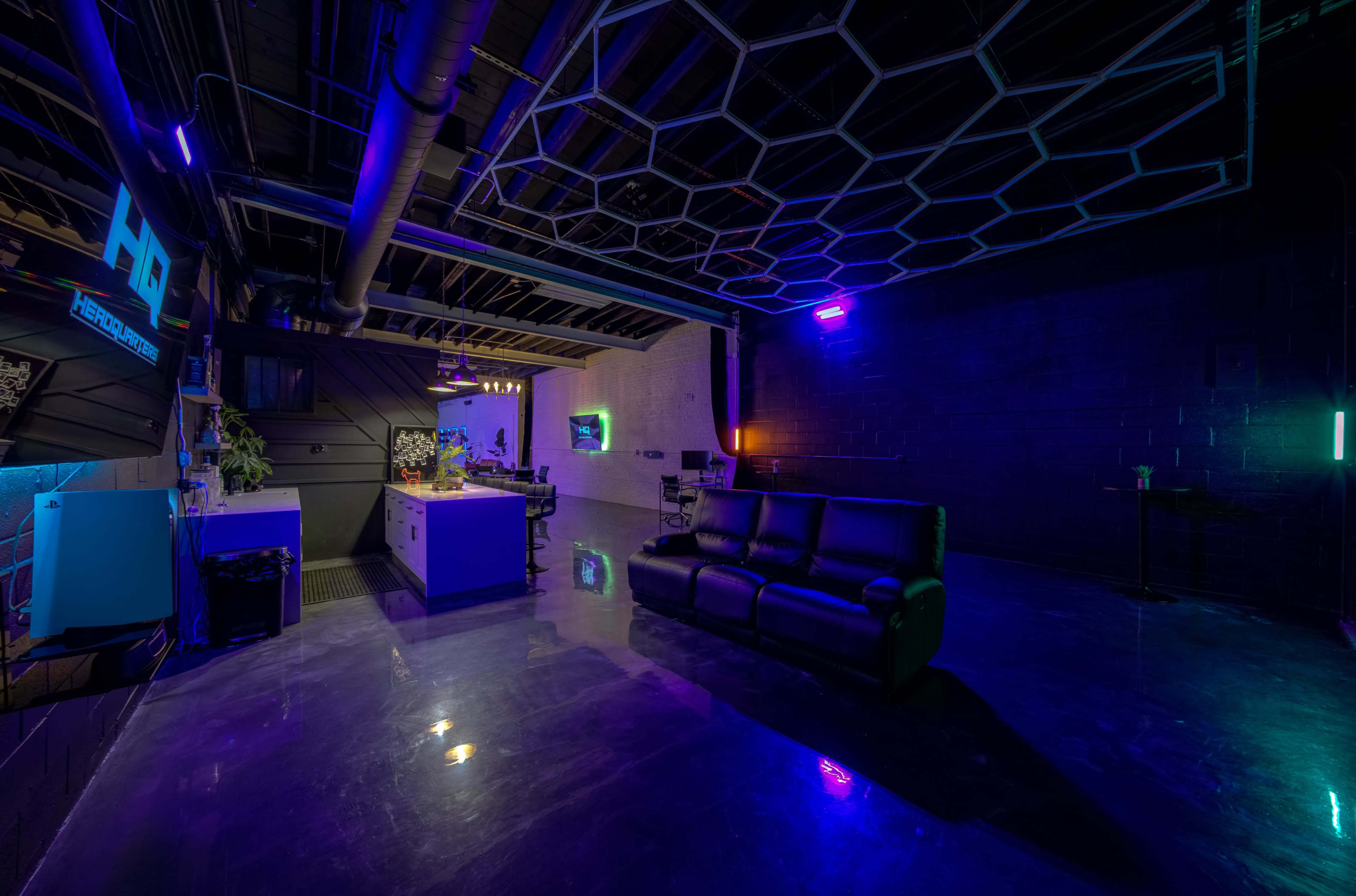 A modern, dimly lit lounge area with a black couch, a sleek kitchen counter, and neon lights casting colors on the walls.