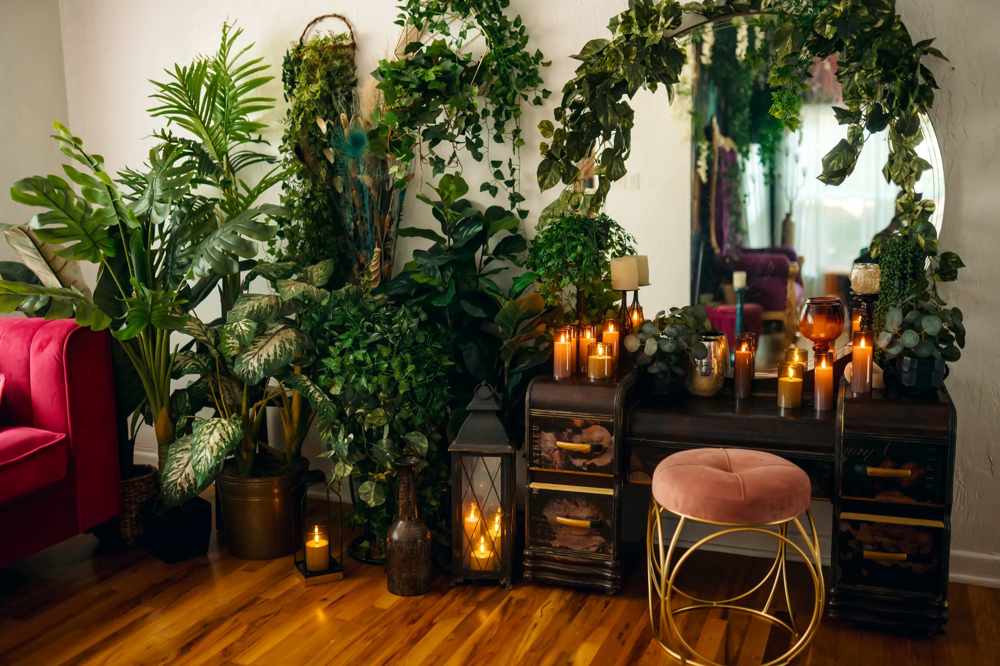 A decorative corner of a room features a mirror surrounded by greenery, candles, and a pink stool alongside a dark wooden dresser.