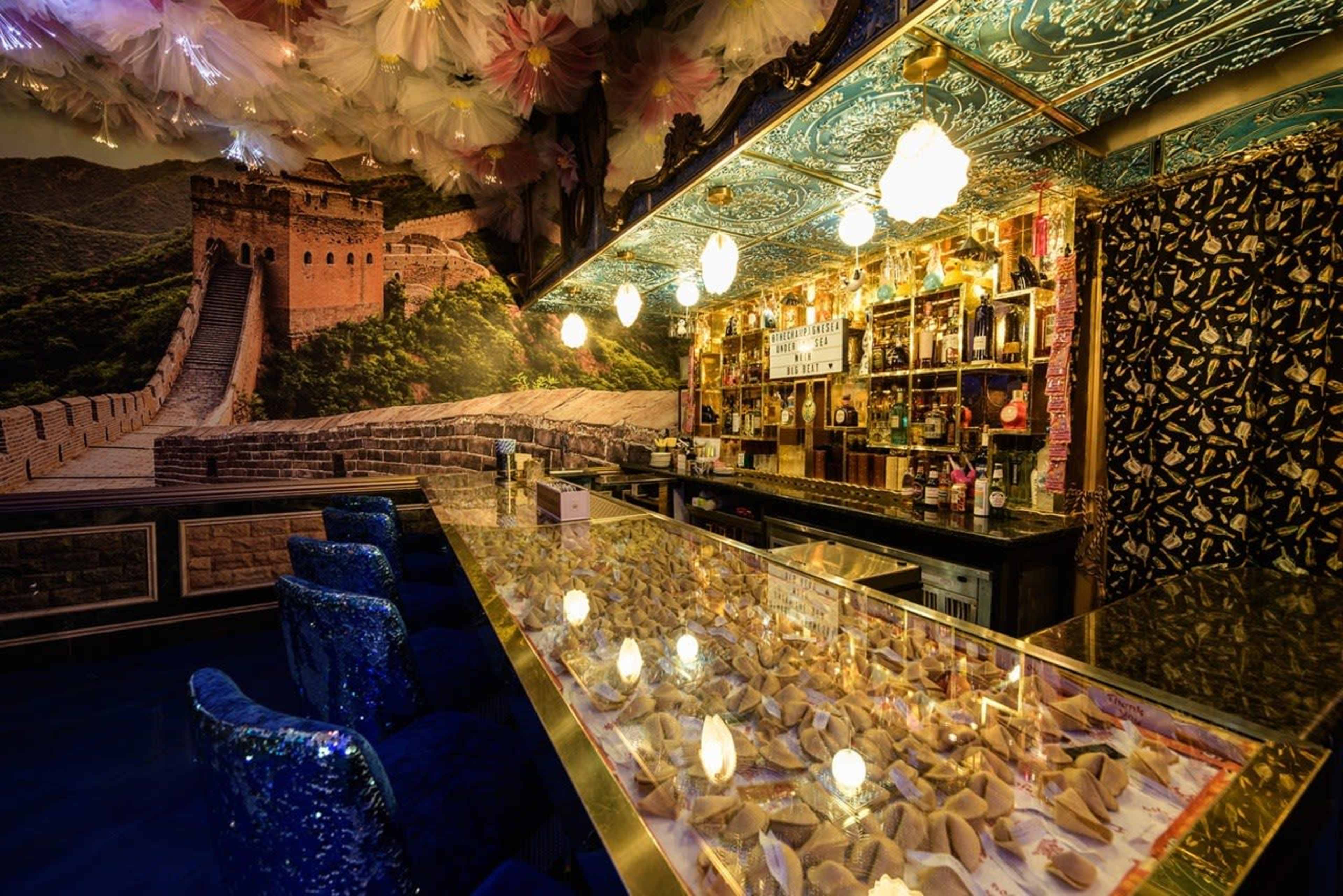 The image shows a bar area with a glass counter showcasing decorative elements, set against a backdrop of the Great Wall of China and equipped with various bottles on shelves.