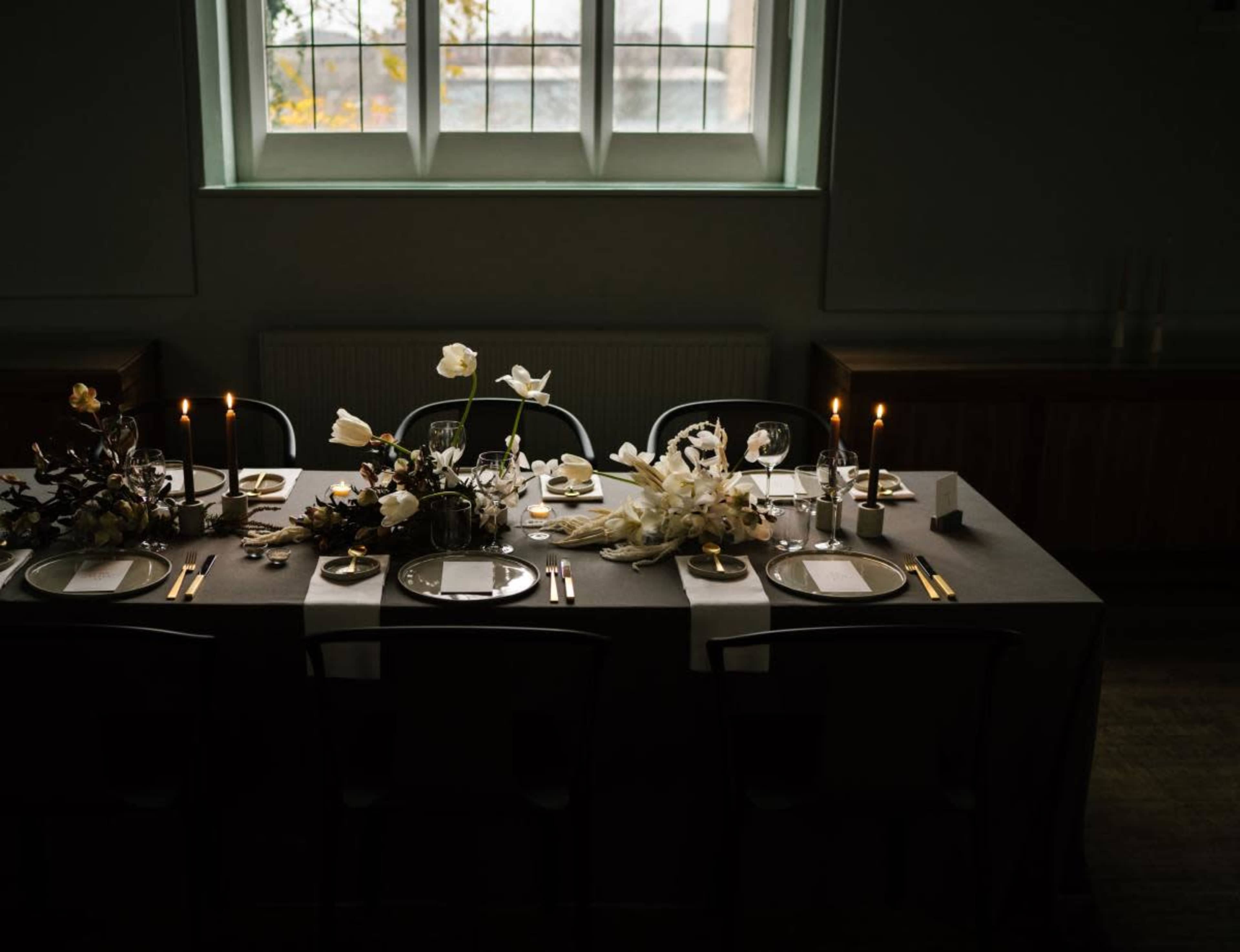 A long dining table is set with candles and floral arrangements in a dimly lit room with a large window.