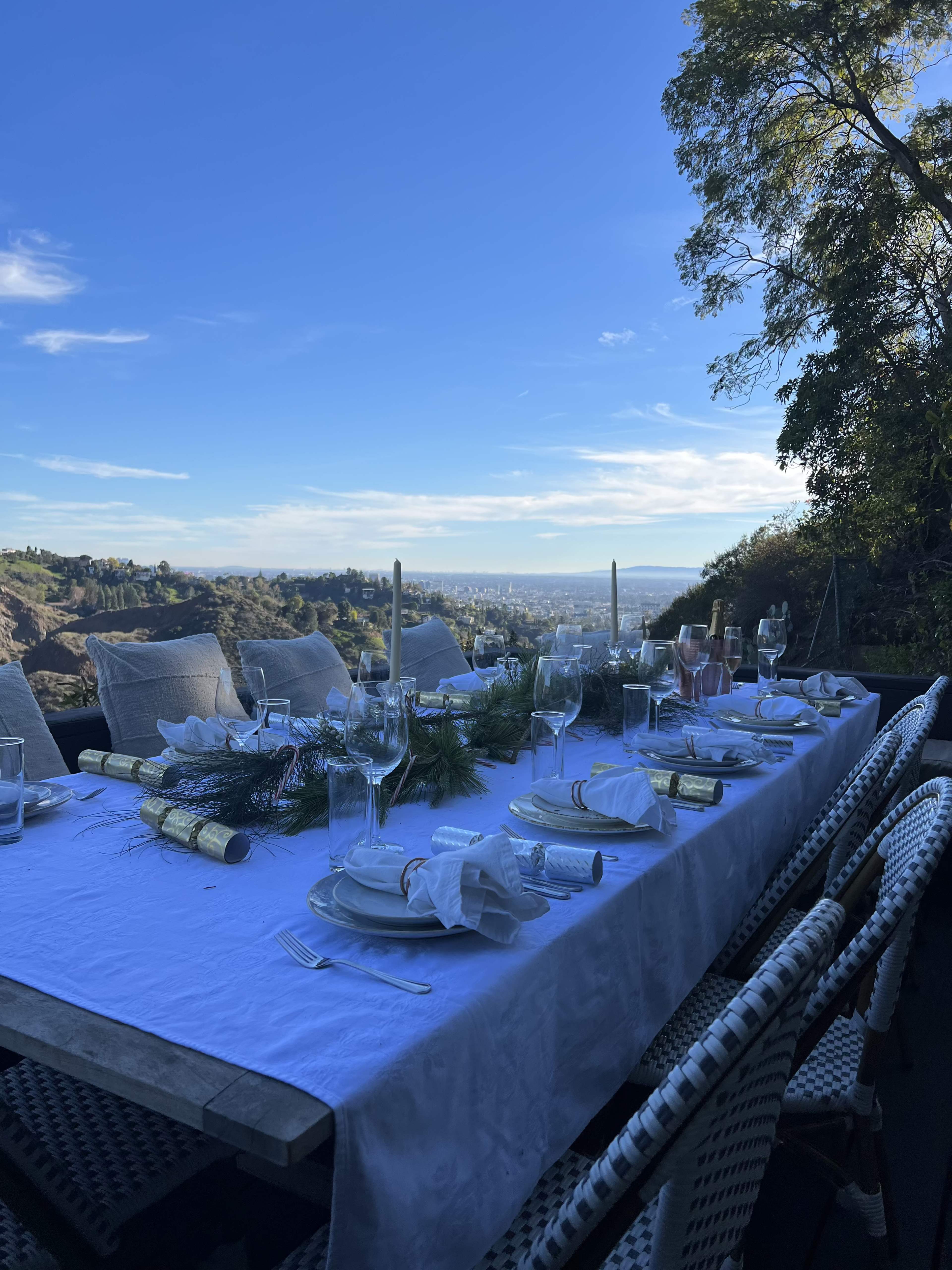 A long outdoor dining table is set with elegant dinnerware, glasses, and festive decorations, overlooking a scenic landscape.