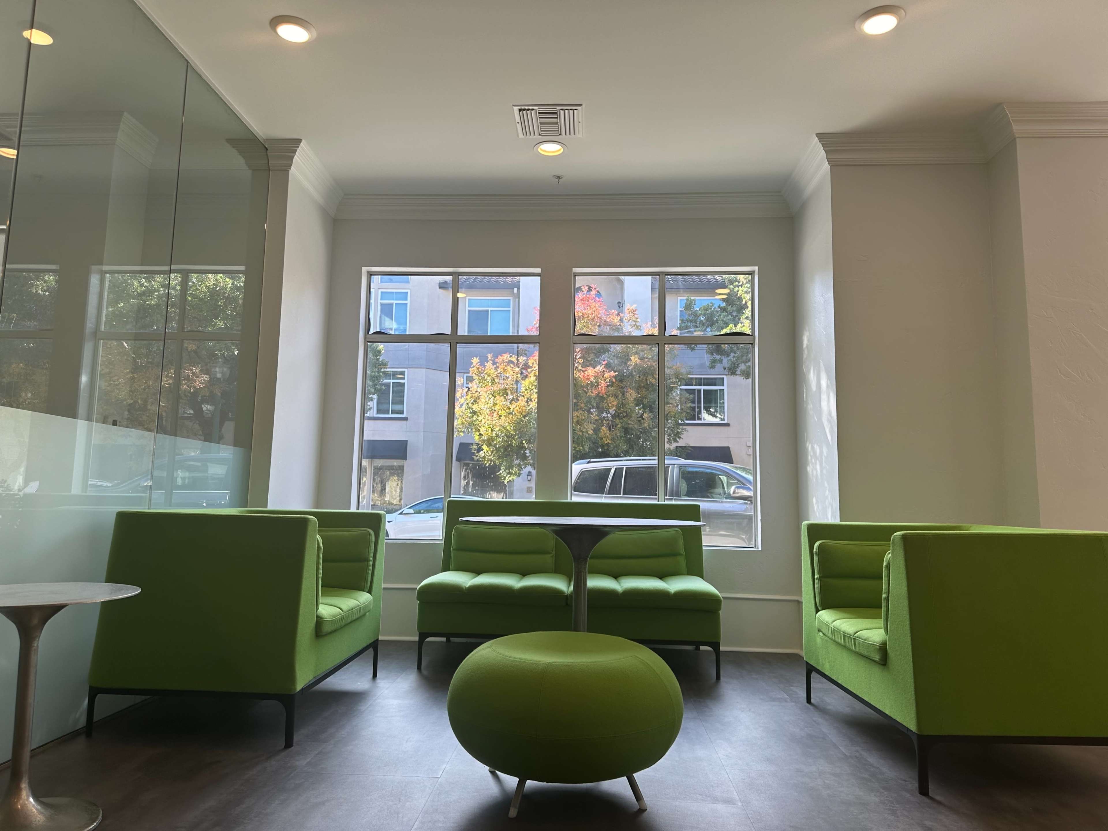 A modern waiting area with green seating, large windows allowing natural light, and a round green table.