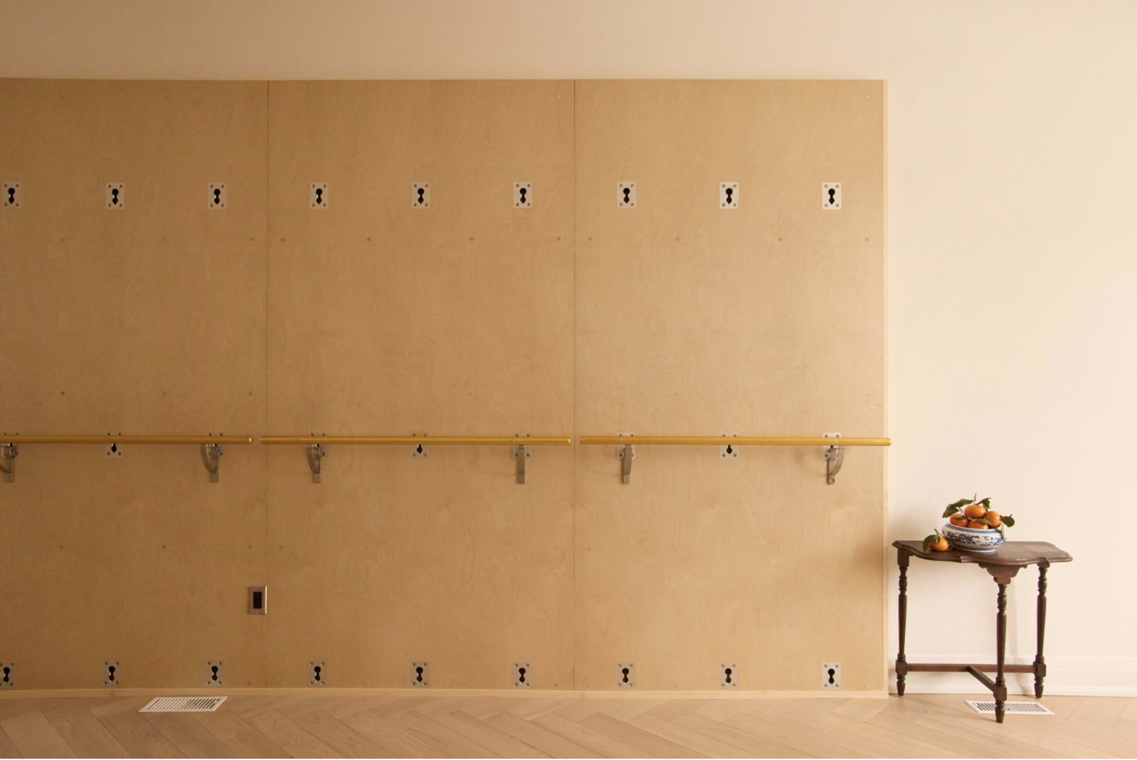 A wooden wall with multiple metal hooks and a yellow strip features a small table with a bowl of fruit next to it.