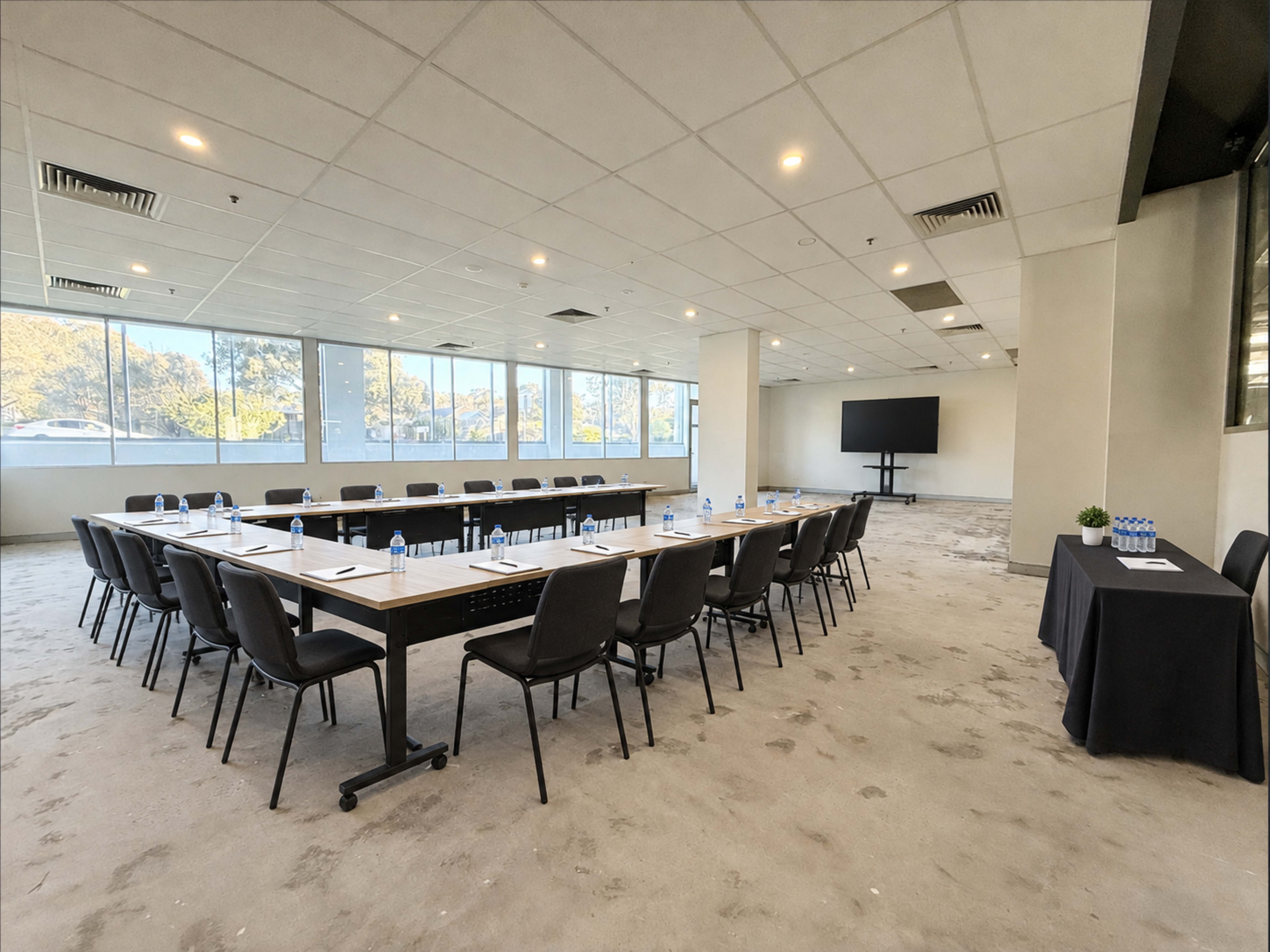 A conference room features a long rectangular table with chairs arranged around it, water bottles, and notepads, alongside a large screen on one wall and windows providing natural light.