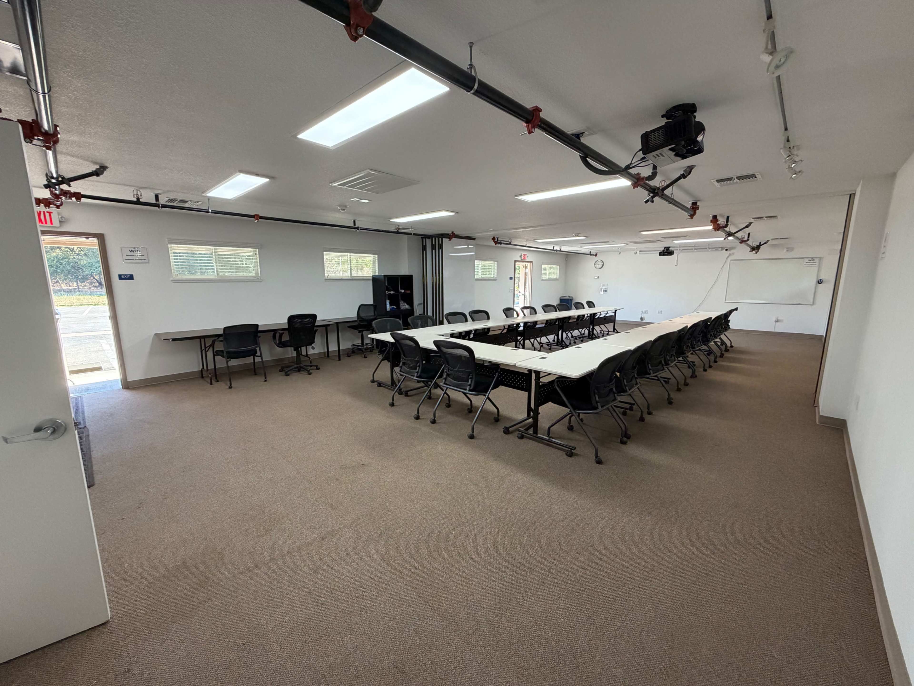 The image shows a spacious meeting room equipped with multiple tables arranged in a U-shape and rows of chairs, along with a projector and fire safety equipment.