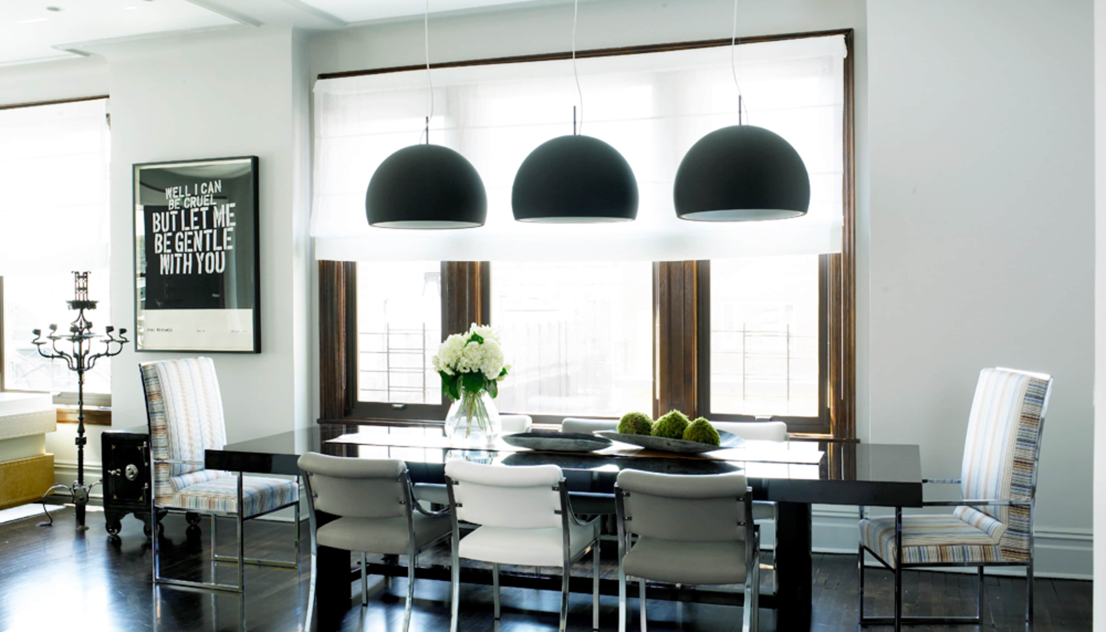 A modern dining room features a long black table surrounded by various chairs, with three pendant lights hanging above and a decorative floral arrangement on the table.