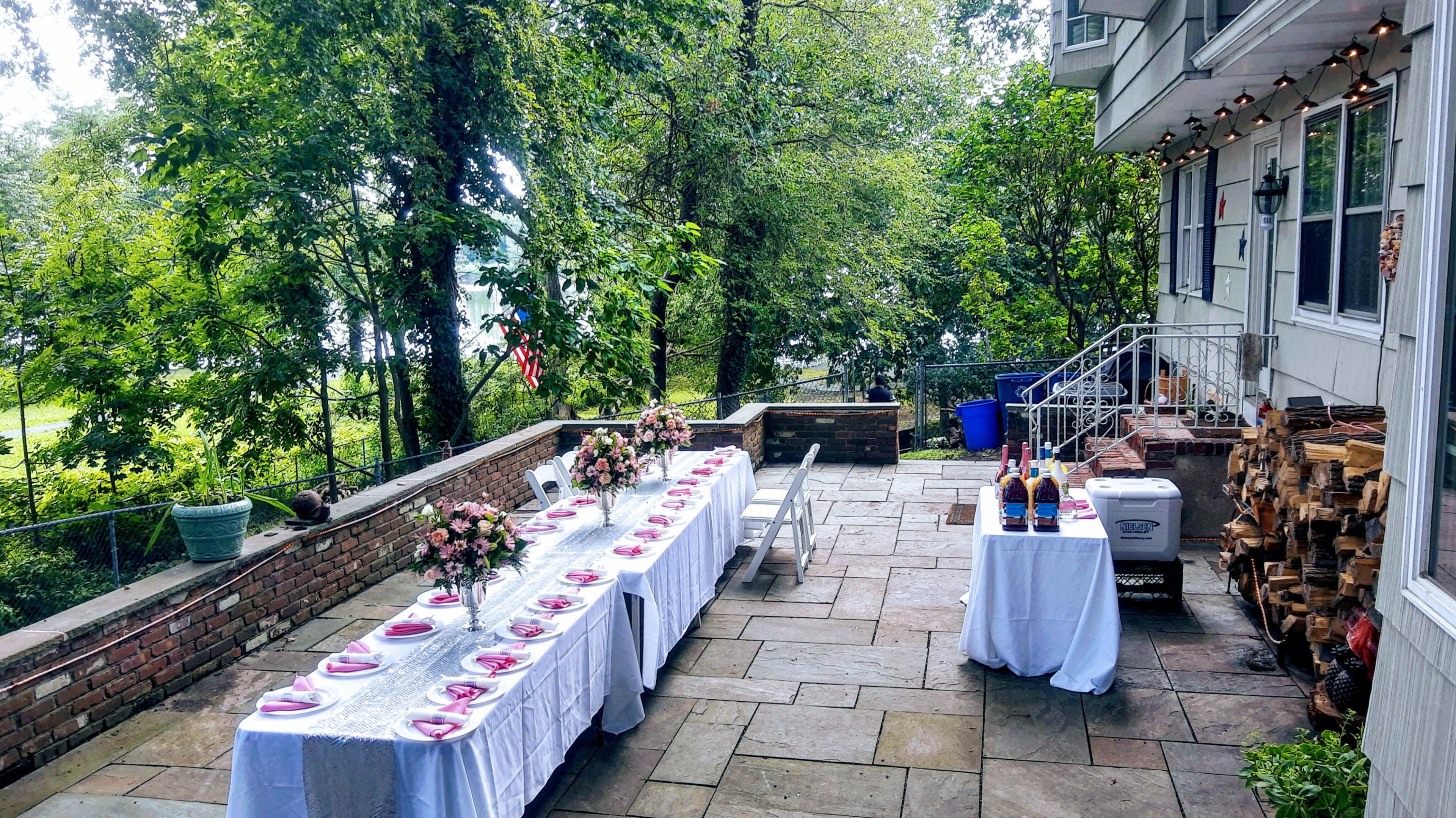 A long table set with pink plates and floral arrangements is positioned on a stone patio surrounded by greenery and a view of the water.