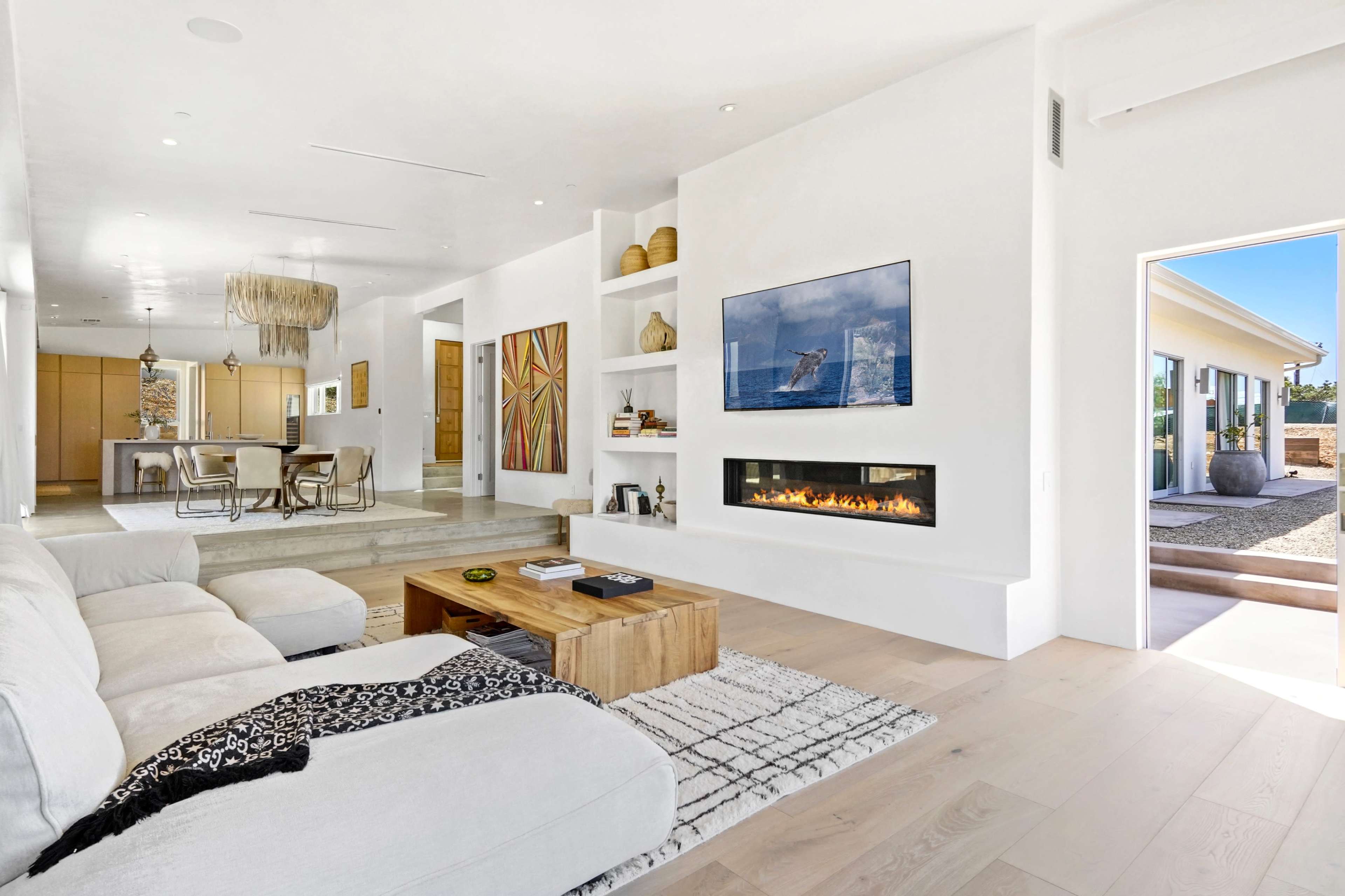 A modern living room features a white sofa, a wooden coffee table, and a wall-mounted TV above a linear fireplace, with an open view to an adjacent kitchen and outdoor space.