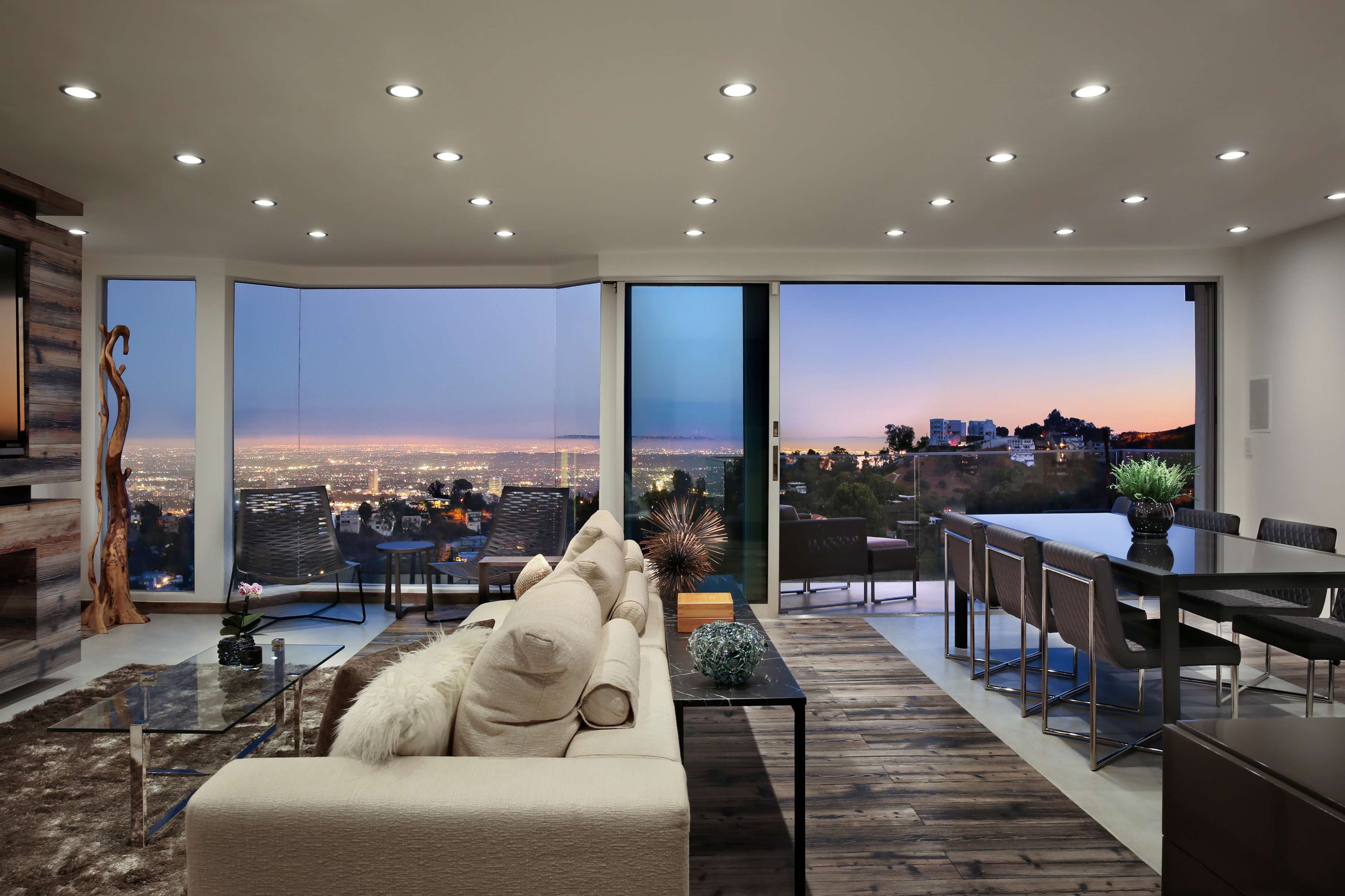A modern living space with large windows offering a panoramic view of a city skyline at sunset.