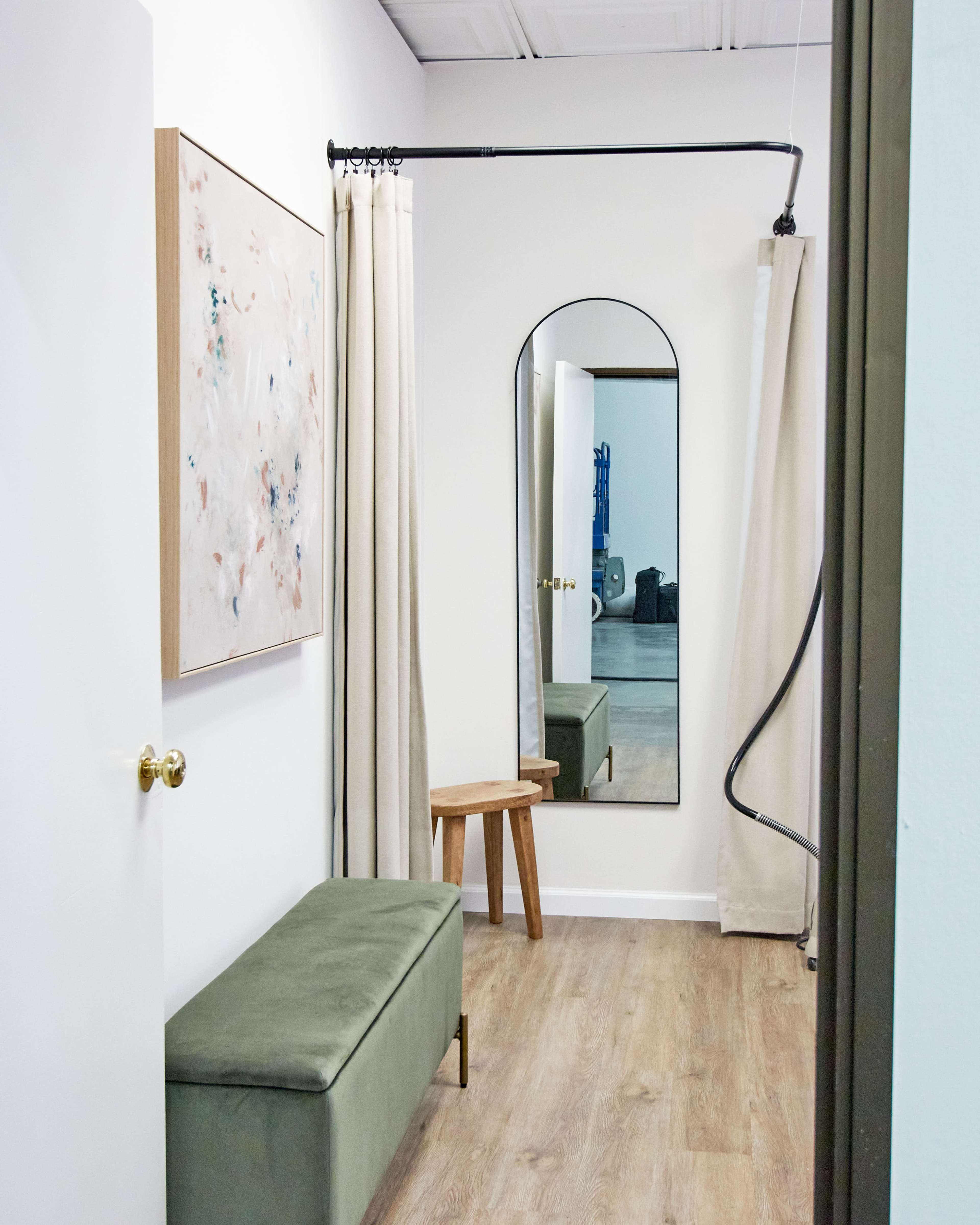A dressing room with a green bench, a full-length mirror with an arched frame, and light-colored curtains.