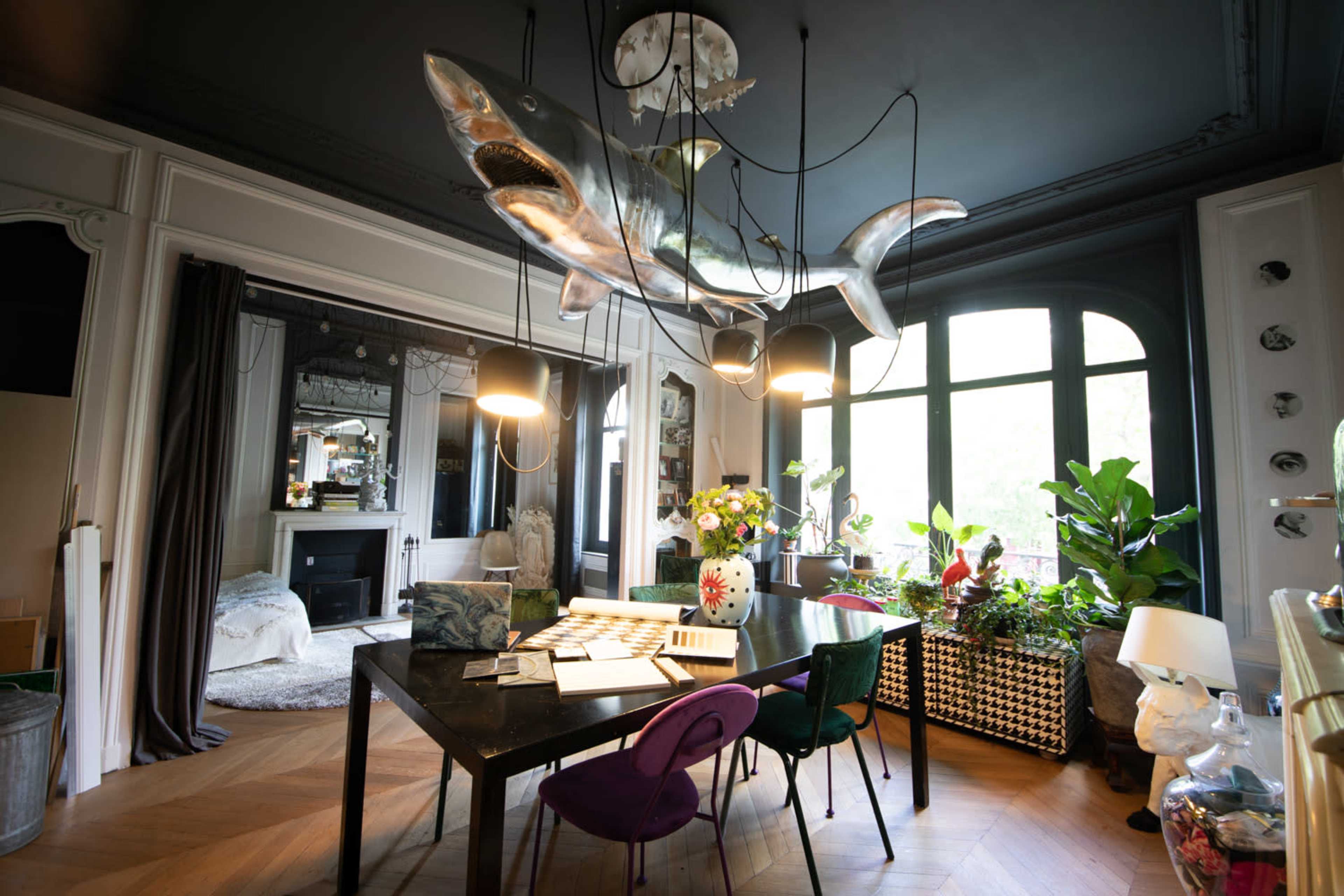 The image shows a dining area with a large, metallic shark sculpture suspended from the ceiling above a black table surrounded by various chairs, with large windows offering natural light.