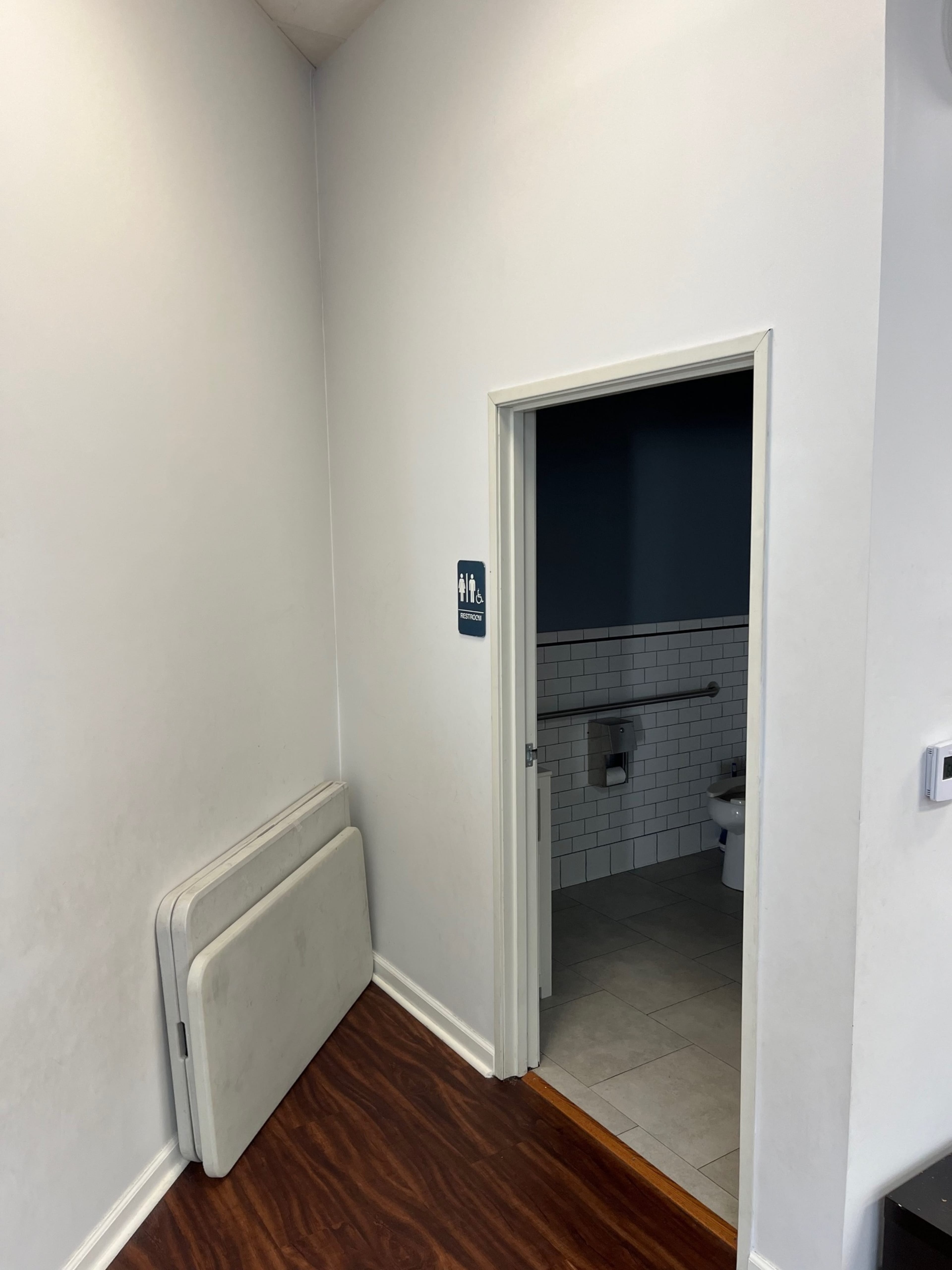 The image shows a corridor with a restroom entrance on the right, featuring a sign indicating it is for both men and women, and two folding tables stacked against the wall.
