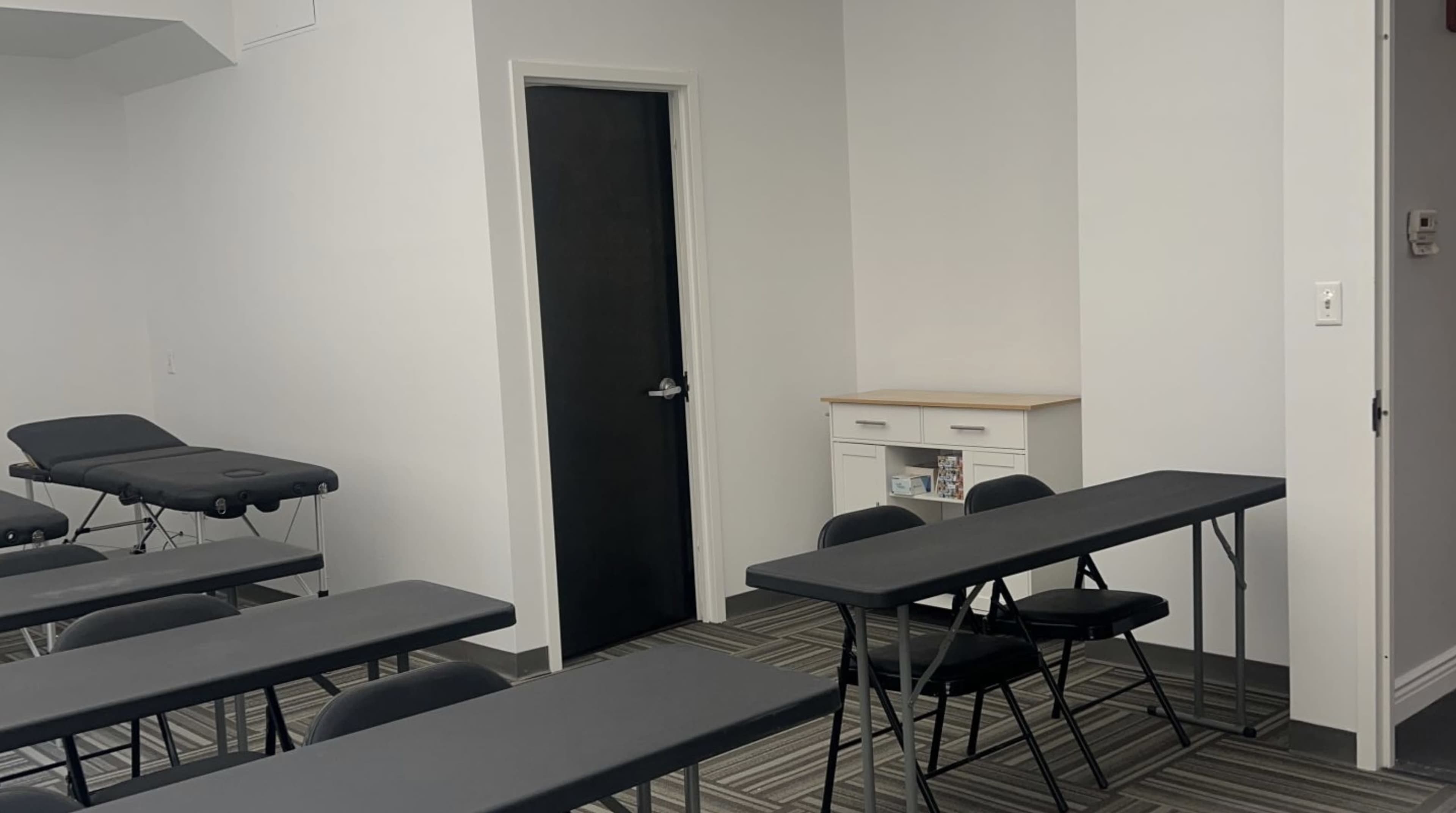 The image shows a room configured with several tables and chairs, a door, and a small cabinet against the wall.