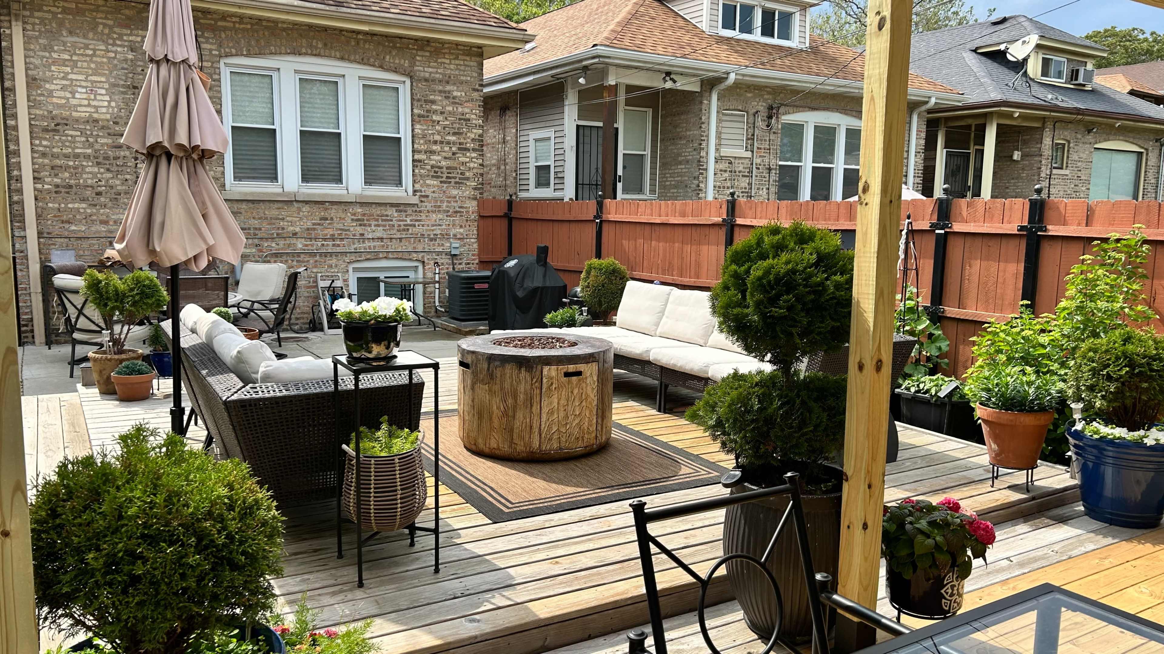 The image shows a backyard patio area featuring a circular fire pit surrounded by seating, planters, and a barbecue grill, with houses in the background and a large umbrella providing shade.