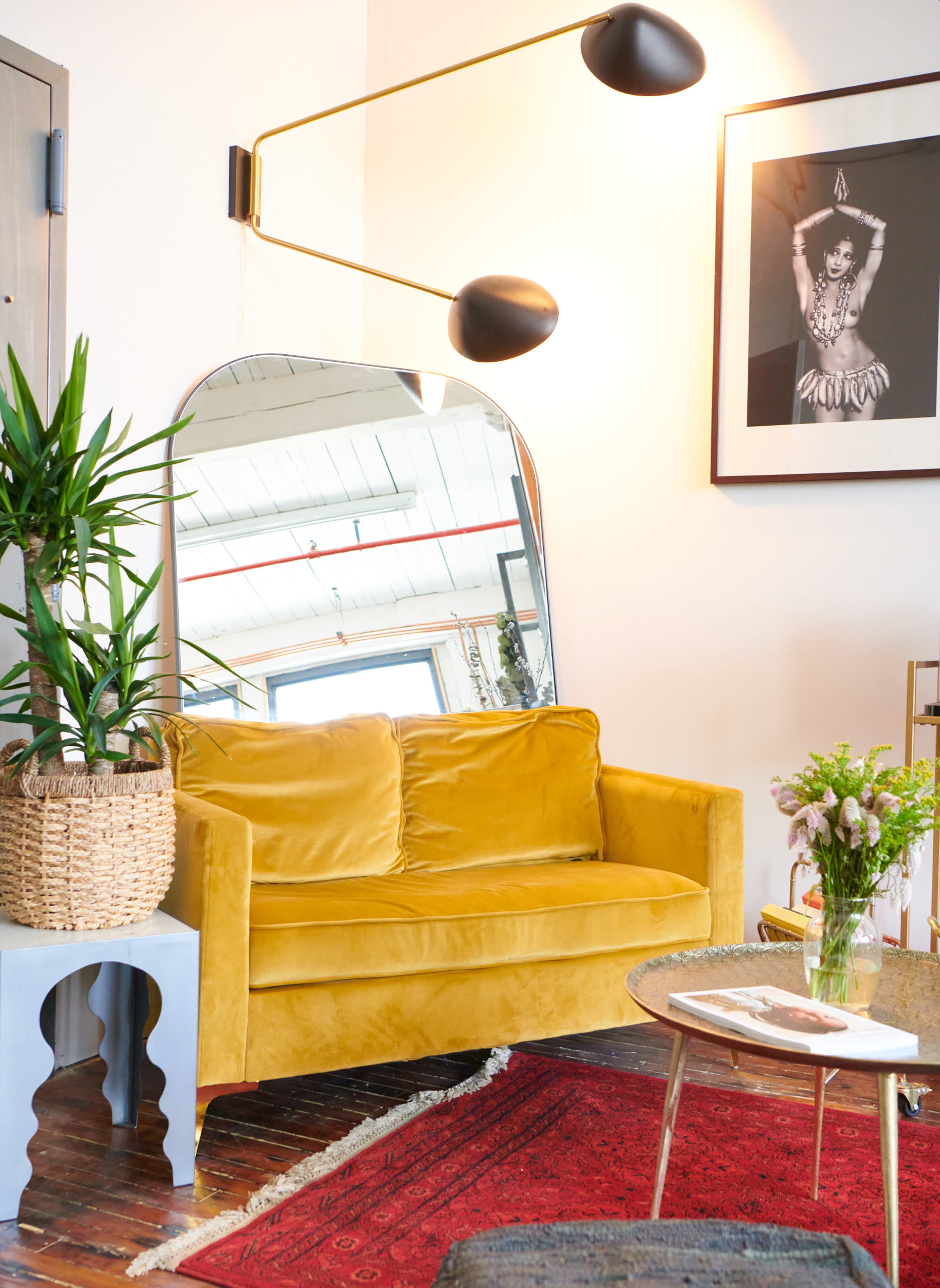 A yellow velvet sofa placed next to a large mirror and a side table, with a potted plant and a floral arrangement in a basket nearby.