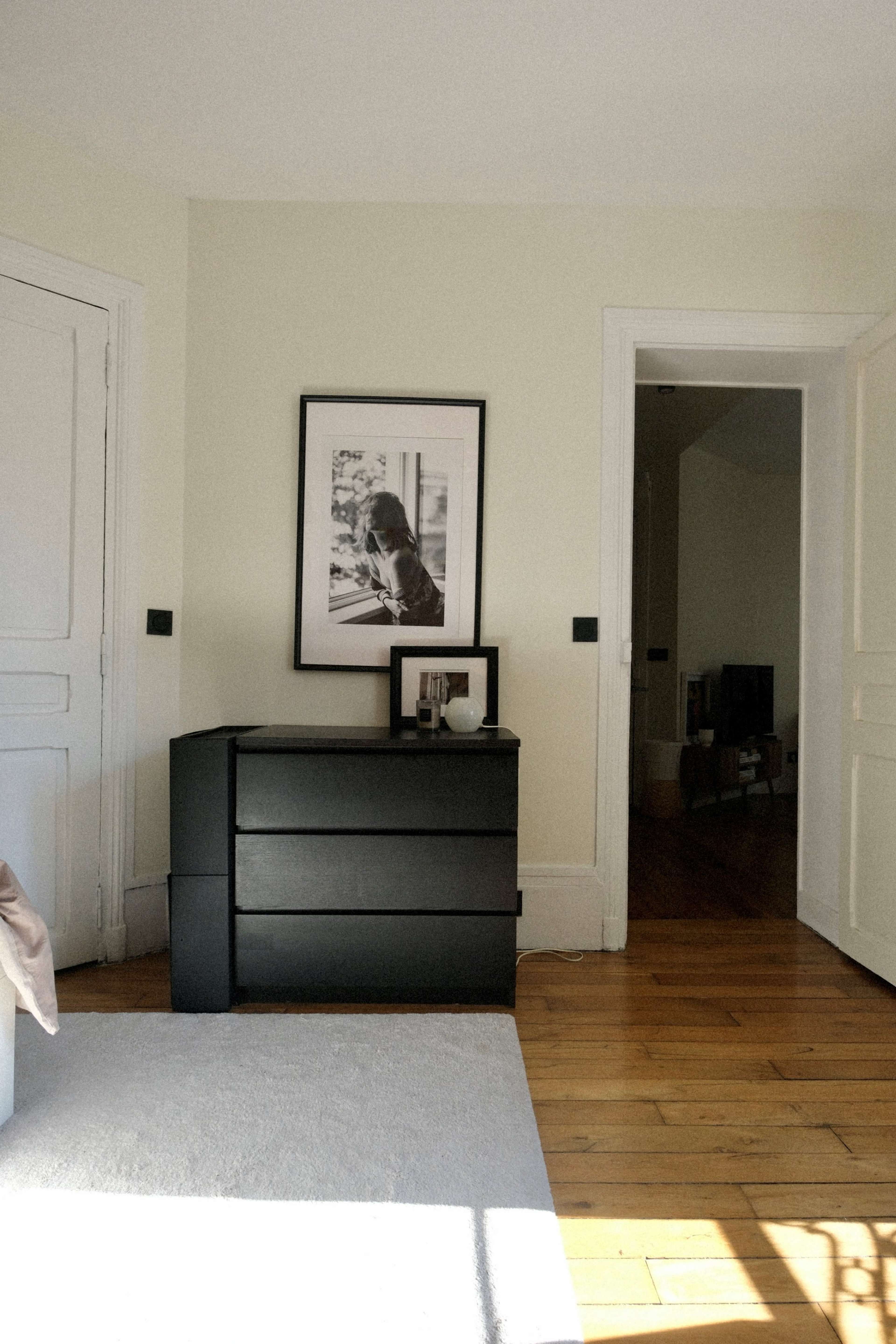 The image shows a minimalist interior with a black dresser, a framed photograph on the wall, and a doorway leading to another room.