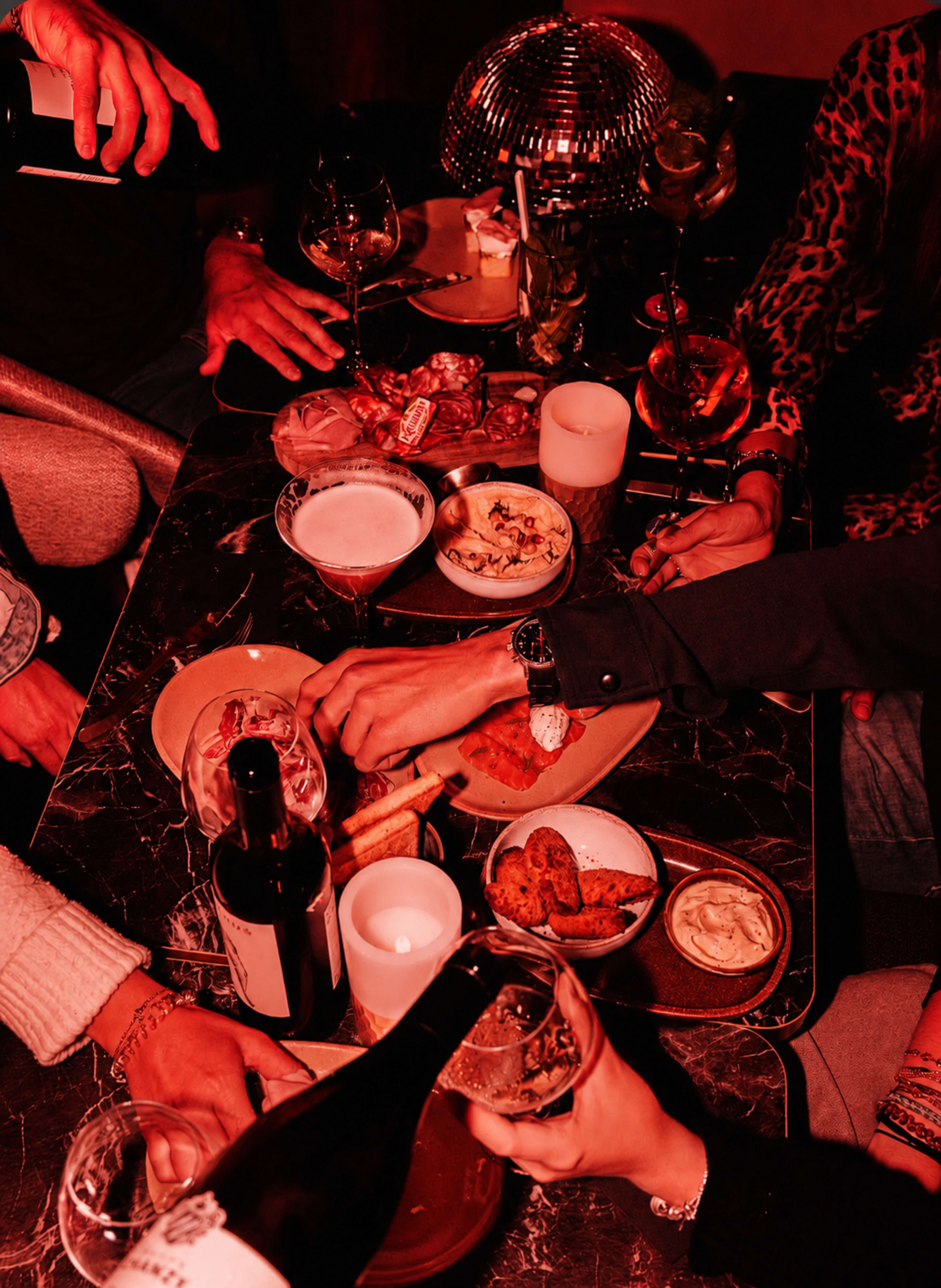A table is filled with various dishes and drinks as several hands reach for food and glasses in a dimly lit setting.
