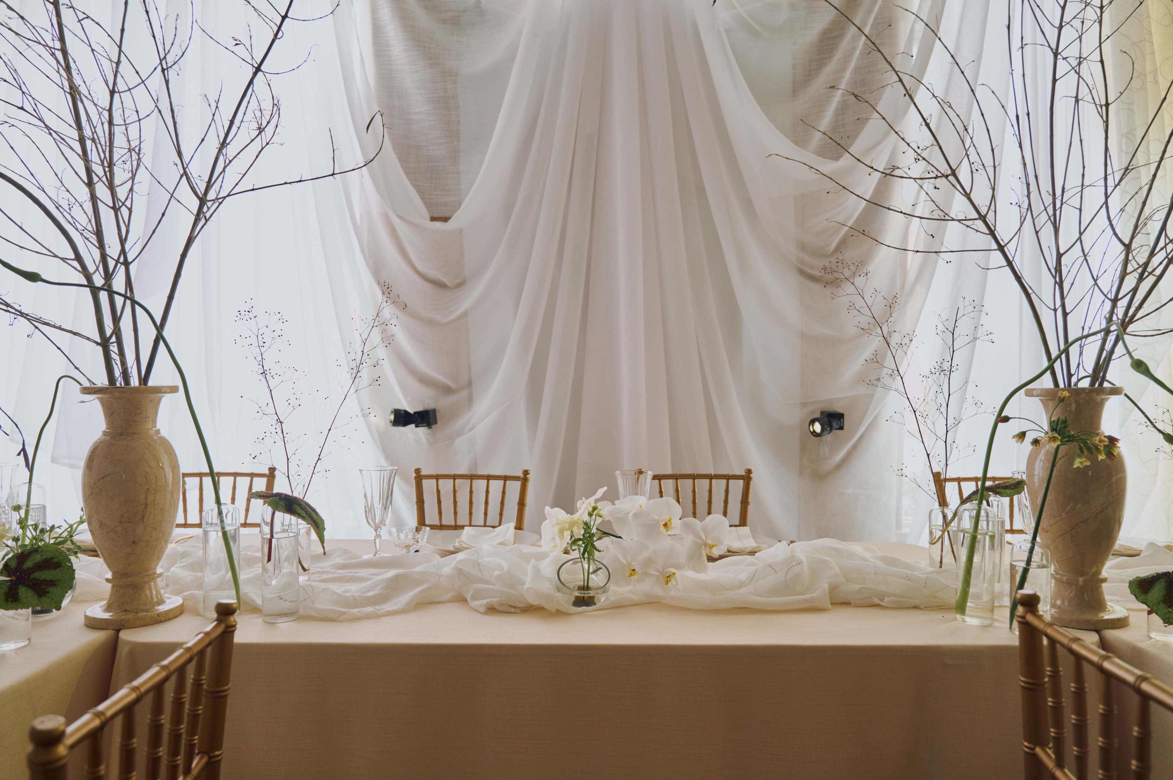 A long banquet table is set with a neutral tablecloth, surrounded by gold chairs, and adorned with decorative branches and white flowers against a backdrop of flowing white fabric.