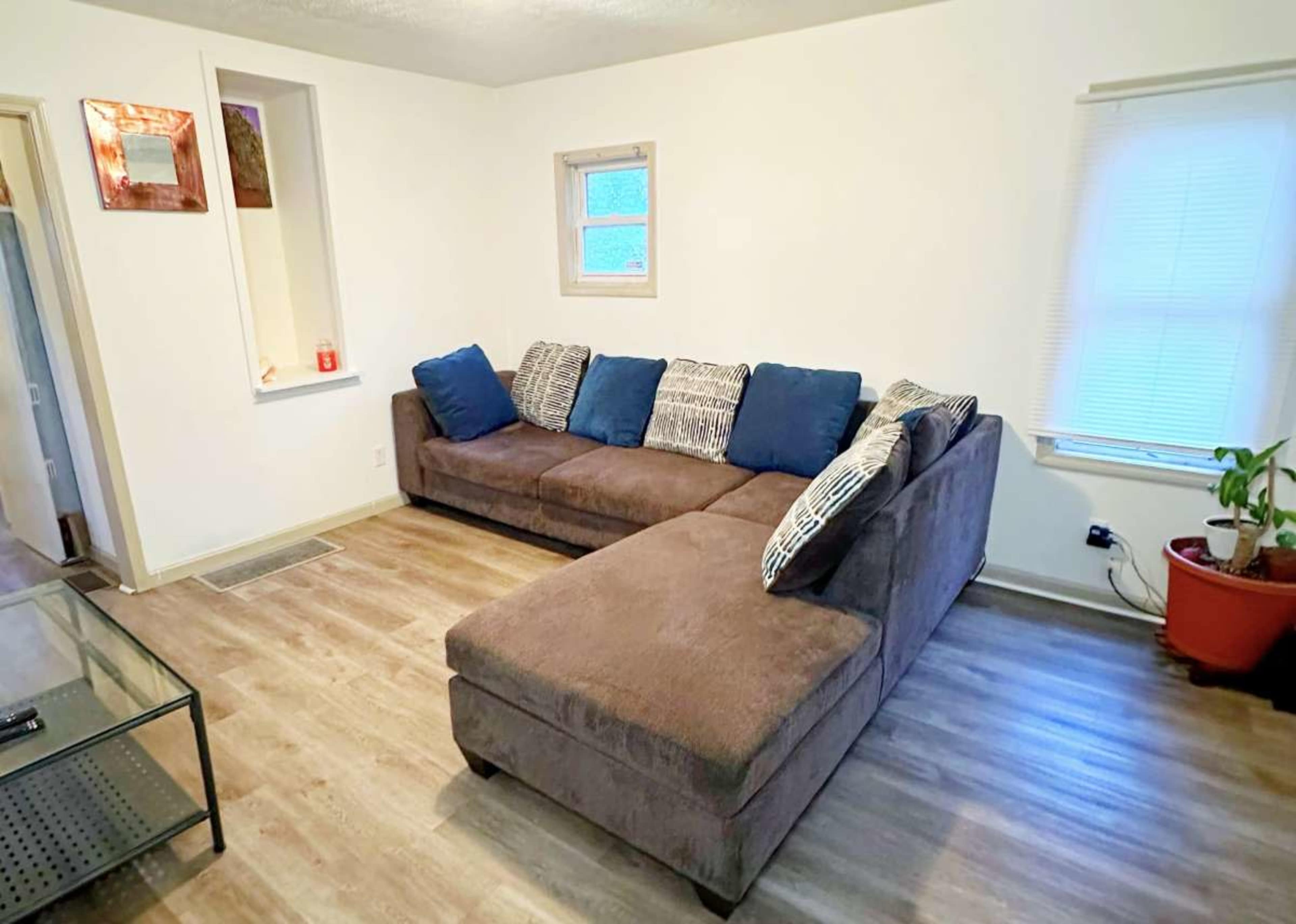 A brown sectional sofa with blue and patterned pillows sits in a bright room featuring a window and a potted plant.