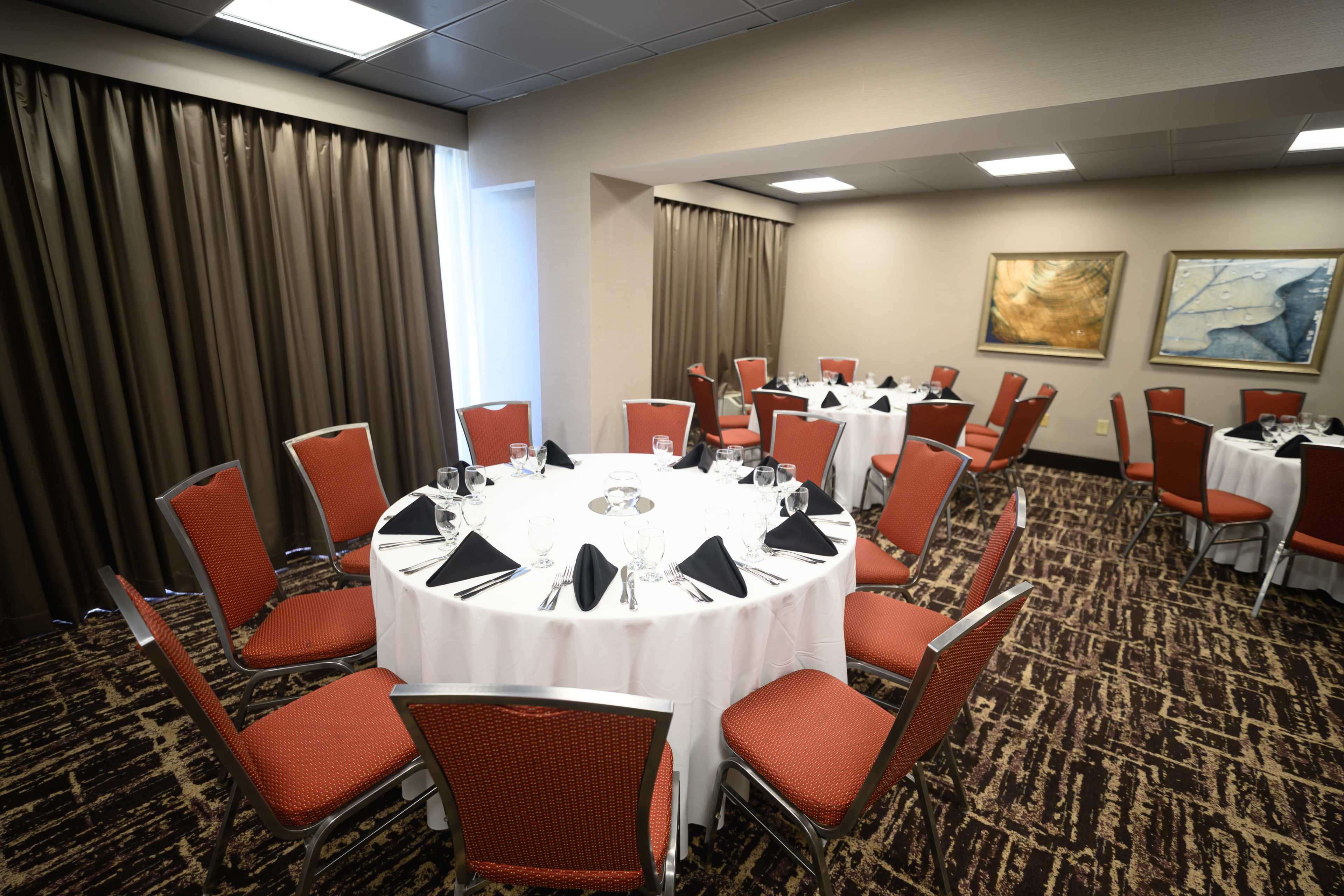 The image shows a banquet room with round tables set for dining, each adorned with white tablecloths, black napkins, and glassware.