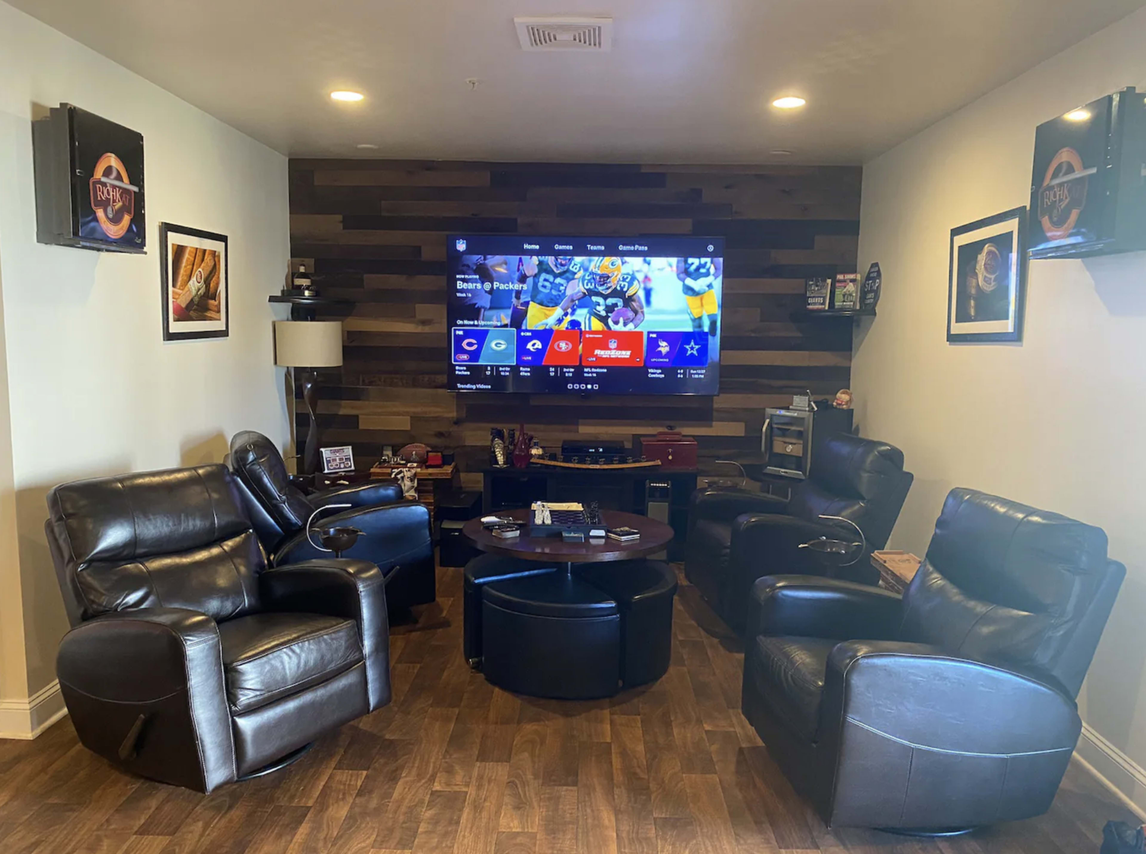 A cozy living room setup with four leather reclining chairs arranged around a circular coffee table, facing a wall-mounted flat-screen TV.
