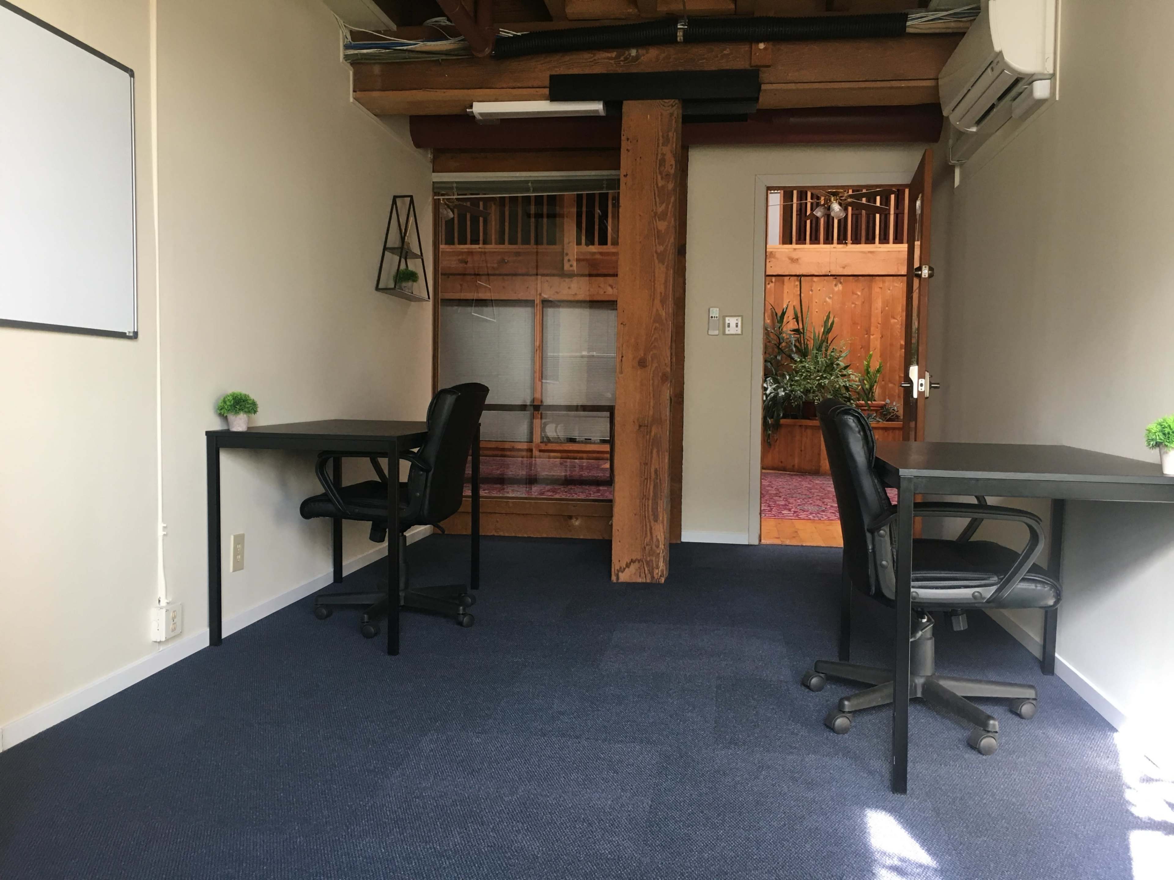 The image shows an office space with two black chairs and desks facing each other, separated by a wooden post in a well-lit room with light-colored walls and a door leading to a brighter area.