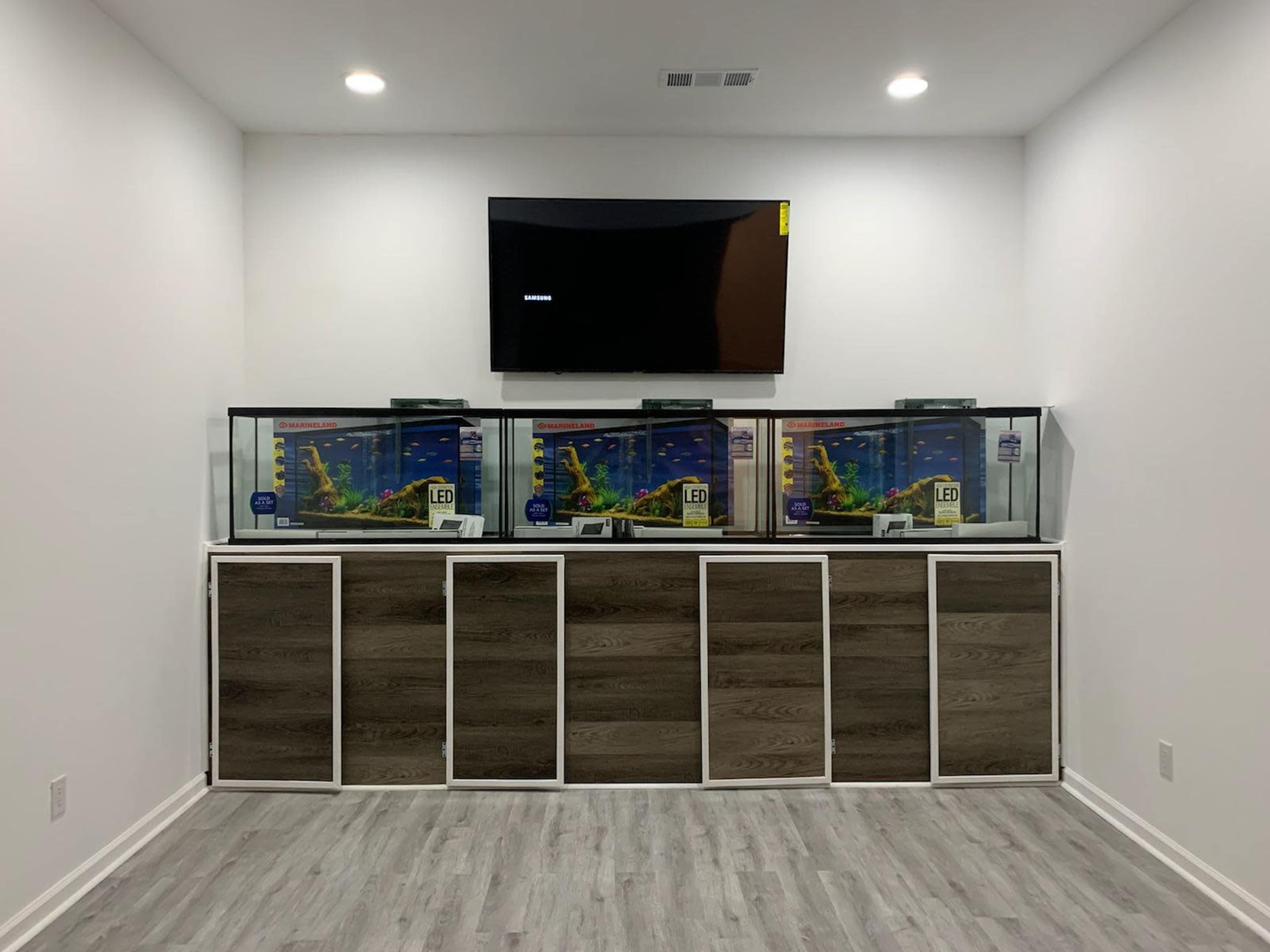 The image shows a room with three illuminated fish tanks placed on a wooden cabinet, accompanied by a flat-screen TV mounted on the wall above.
