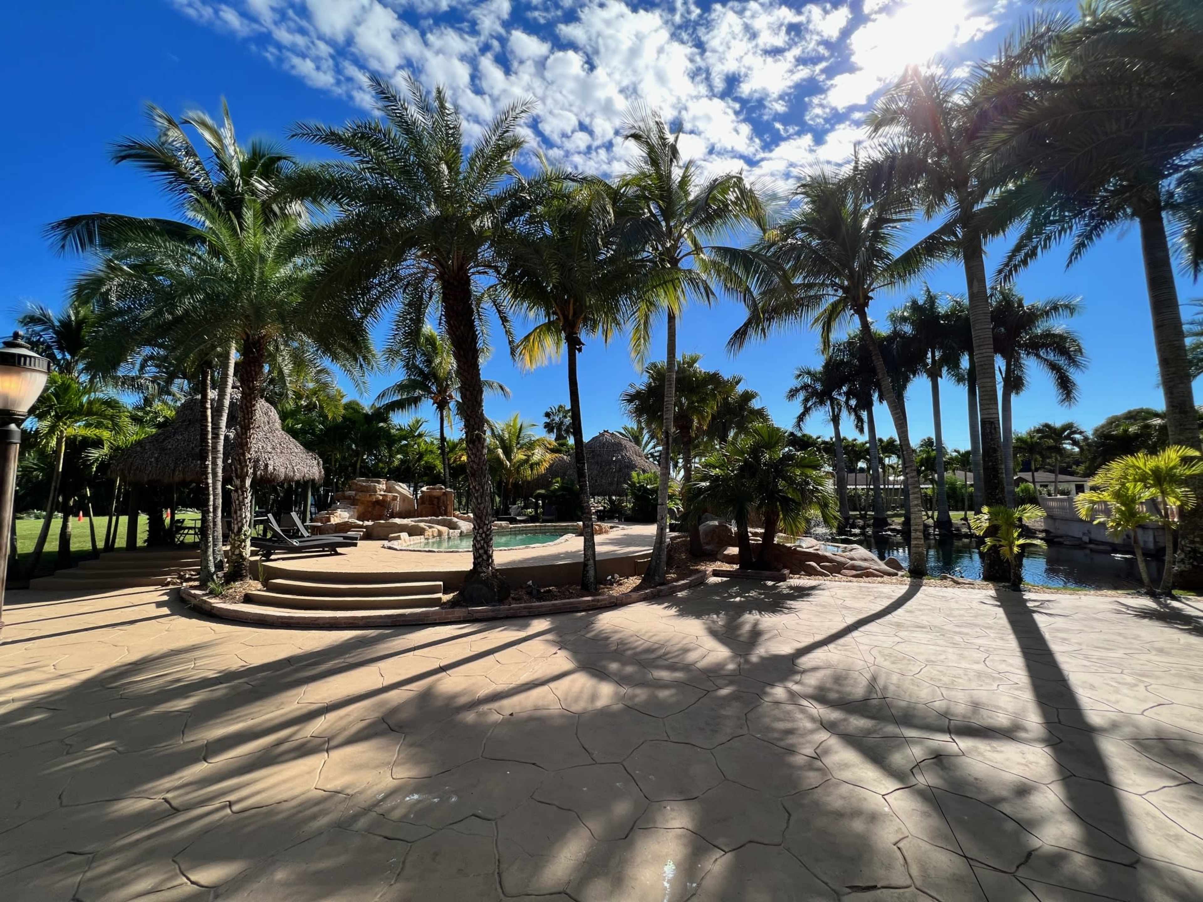 A tropical landscape featuring palm trees, a pool area with a thatched-roof structure, and a clear blue sky.