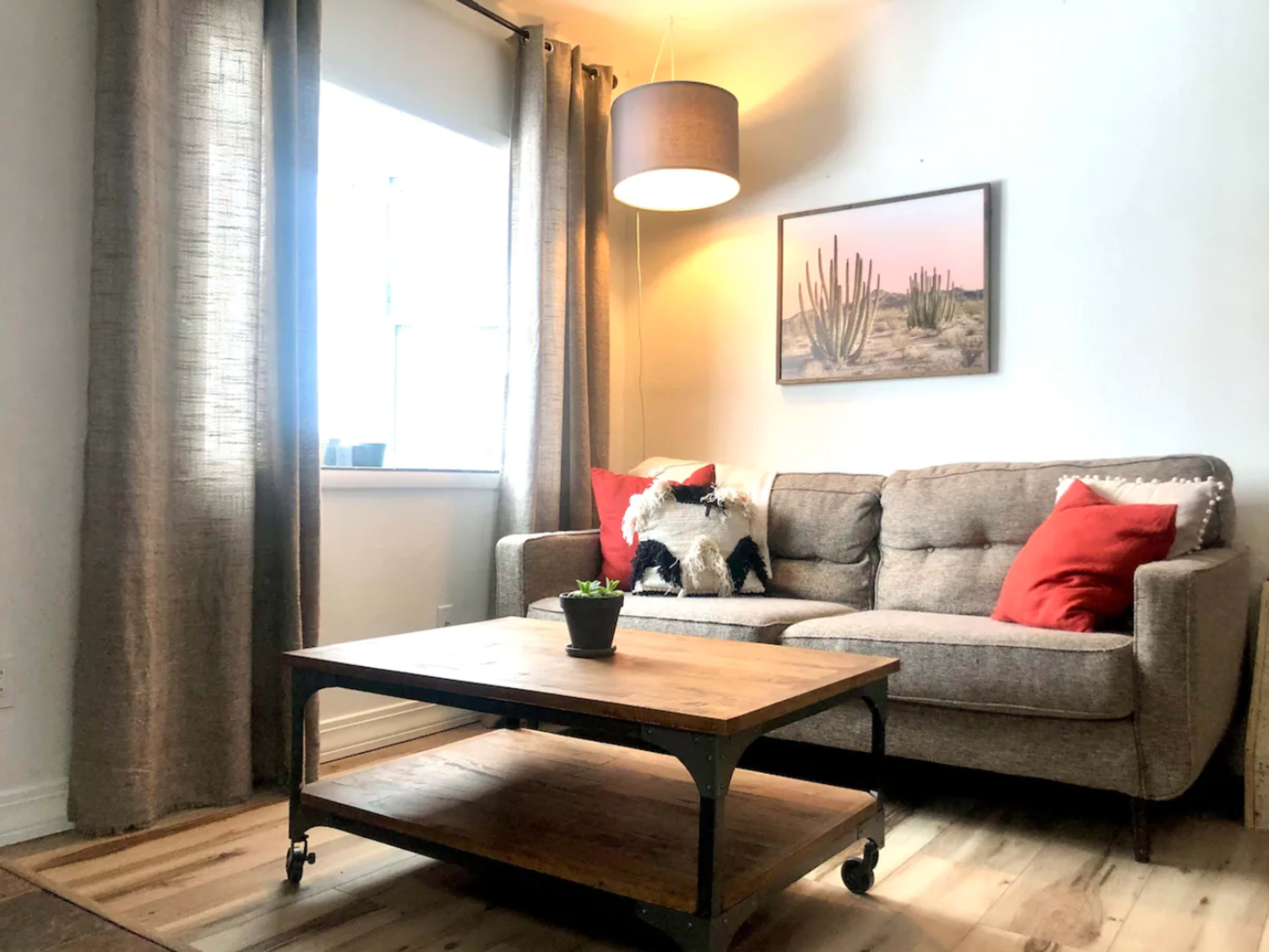 A living room features a gray sofa with red pillows, a wooden coffee table, and a large window adorned with curtains, illuminated by a hanging light.