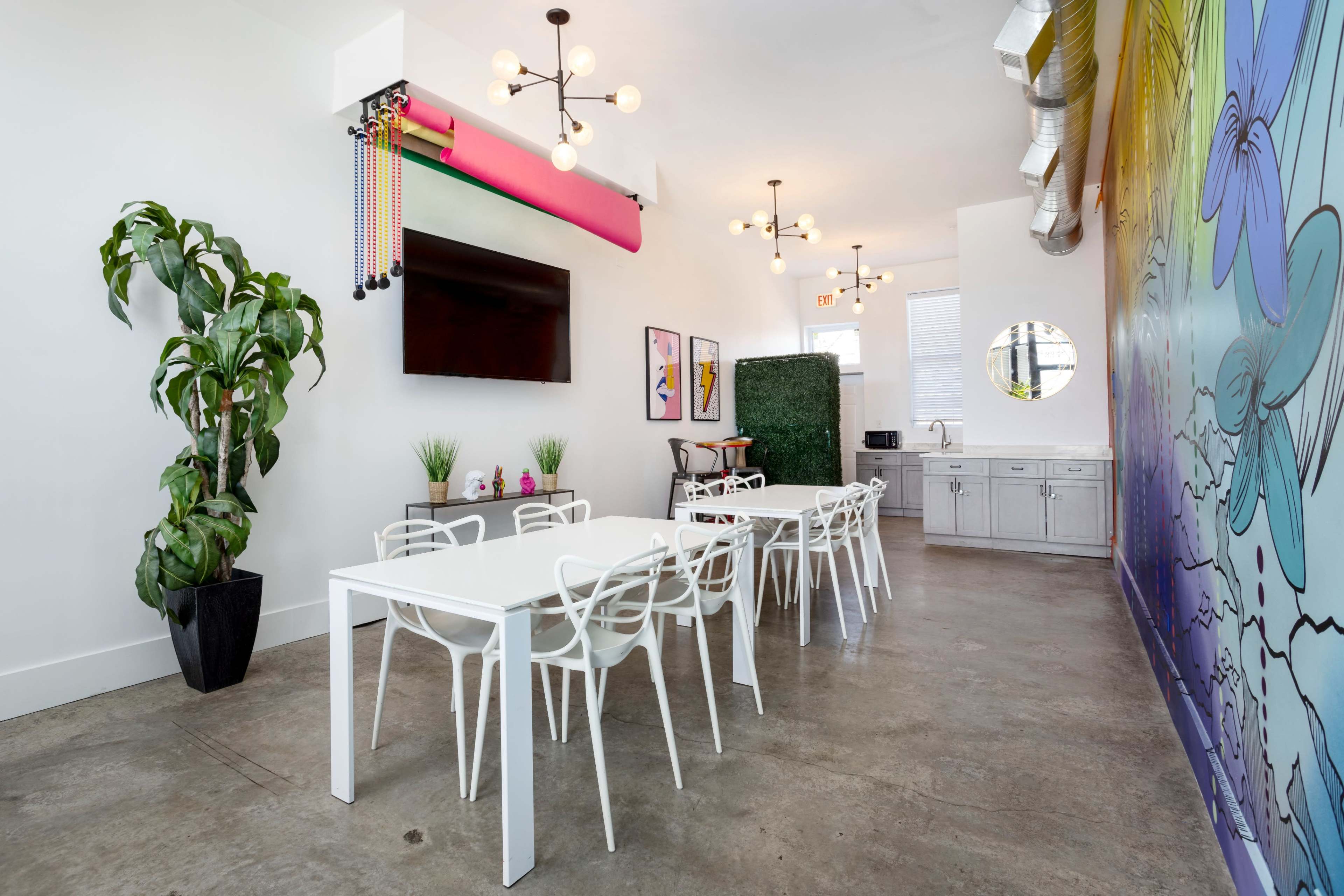 The image shows a bright, modern interior with white tables and chairs, a large wall mural, and decorative plants.