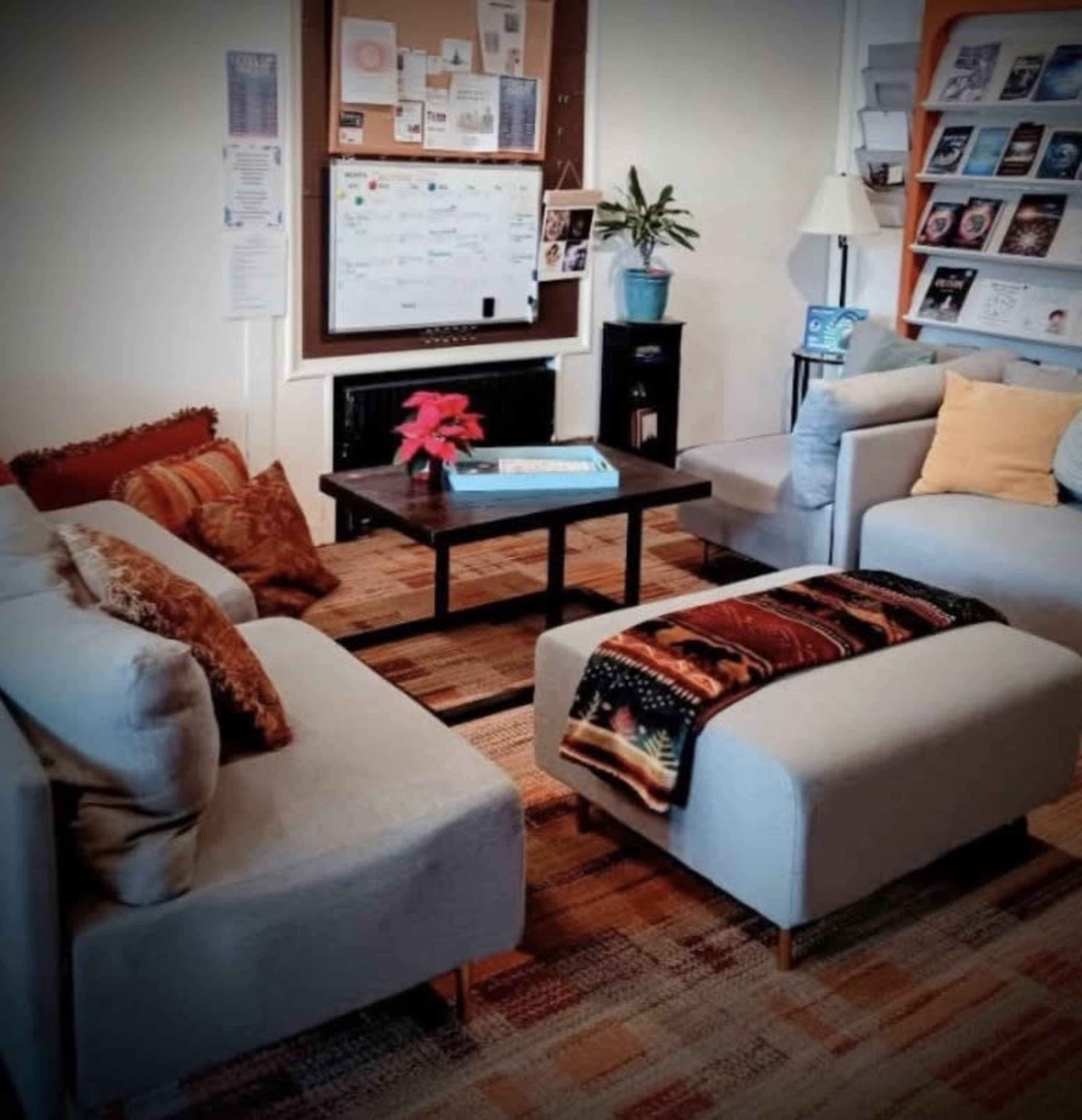 The image shows a cozy lounge area with two light gray sofas, a wooden coffee table, and a bookshelf filled with books, complemented by a potted plant and a bulletin board on the wall.
