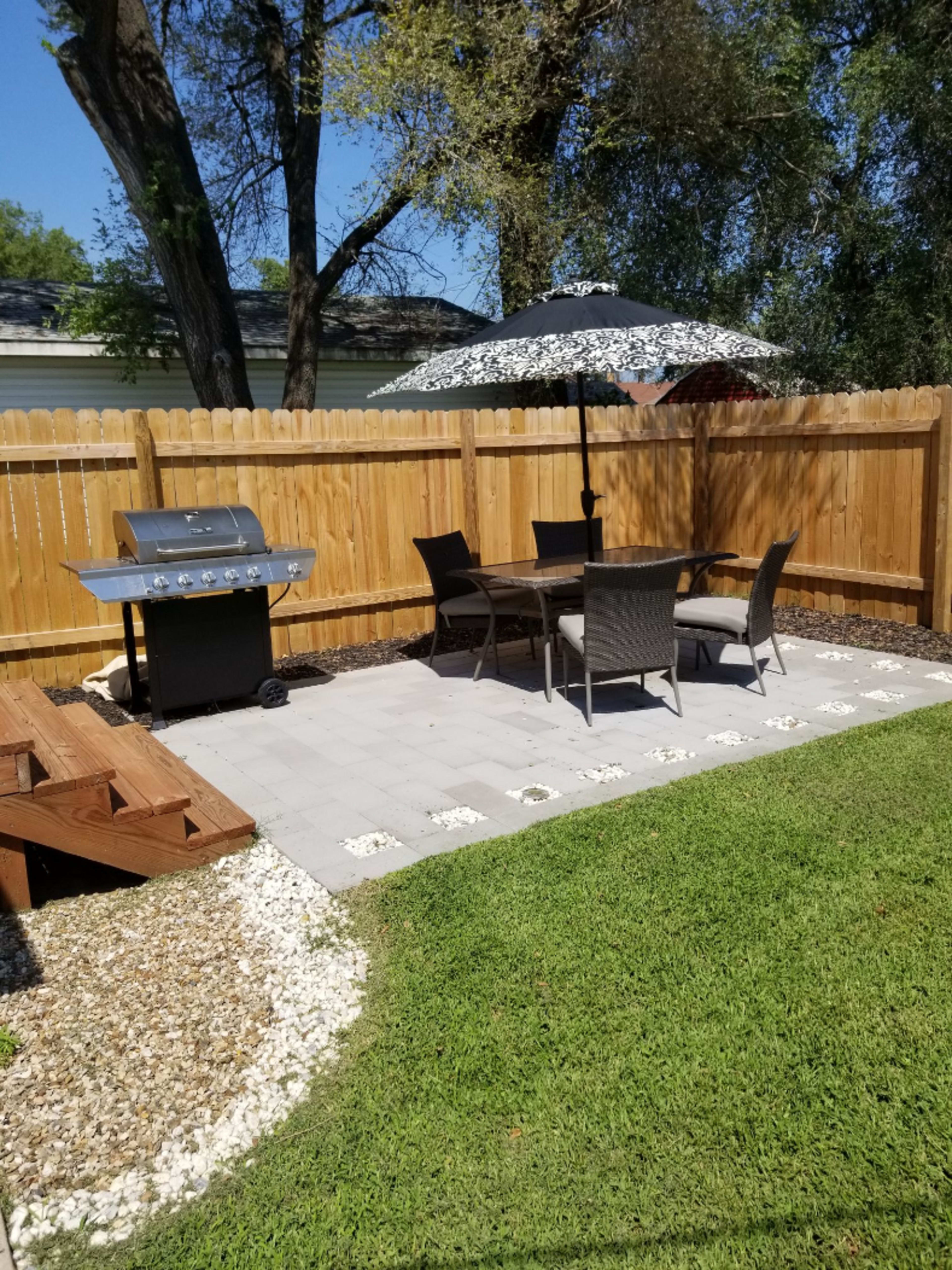 A backyard patio features a grill, dining table with chairs, and an umbrella, surrounded by a wooden fence and landscaped with stones and grass.