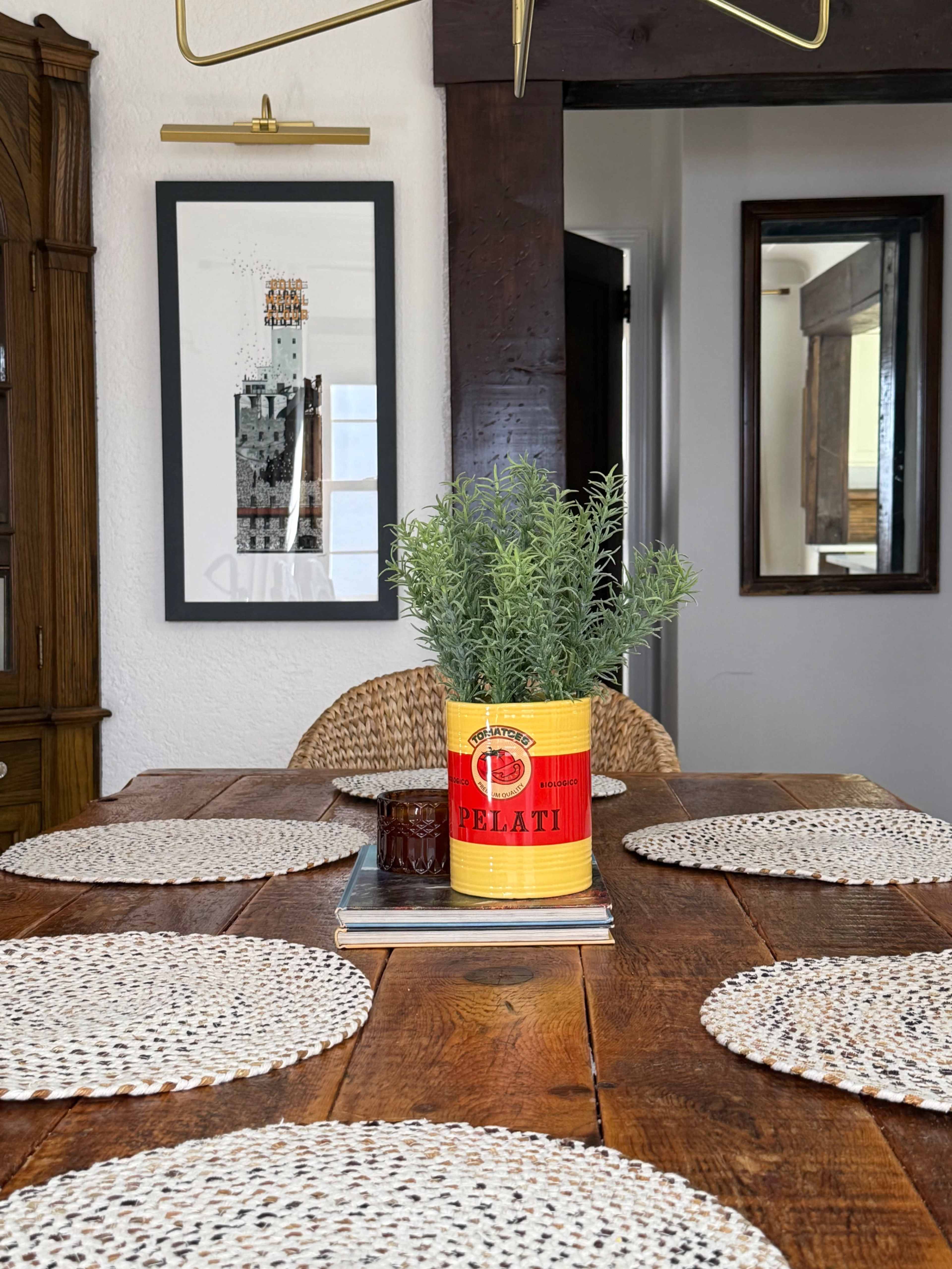 A dining table set with woven placemats, a vintage red can containing herbs, and framed art on the walls.