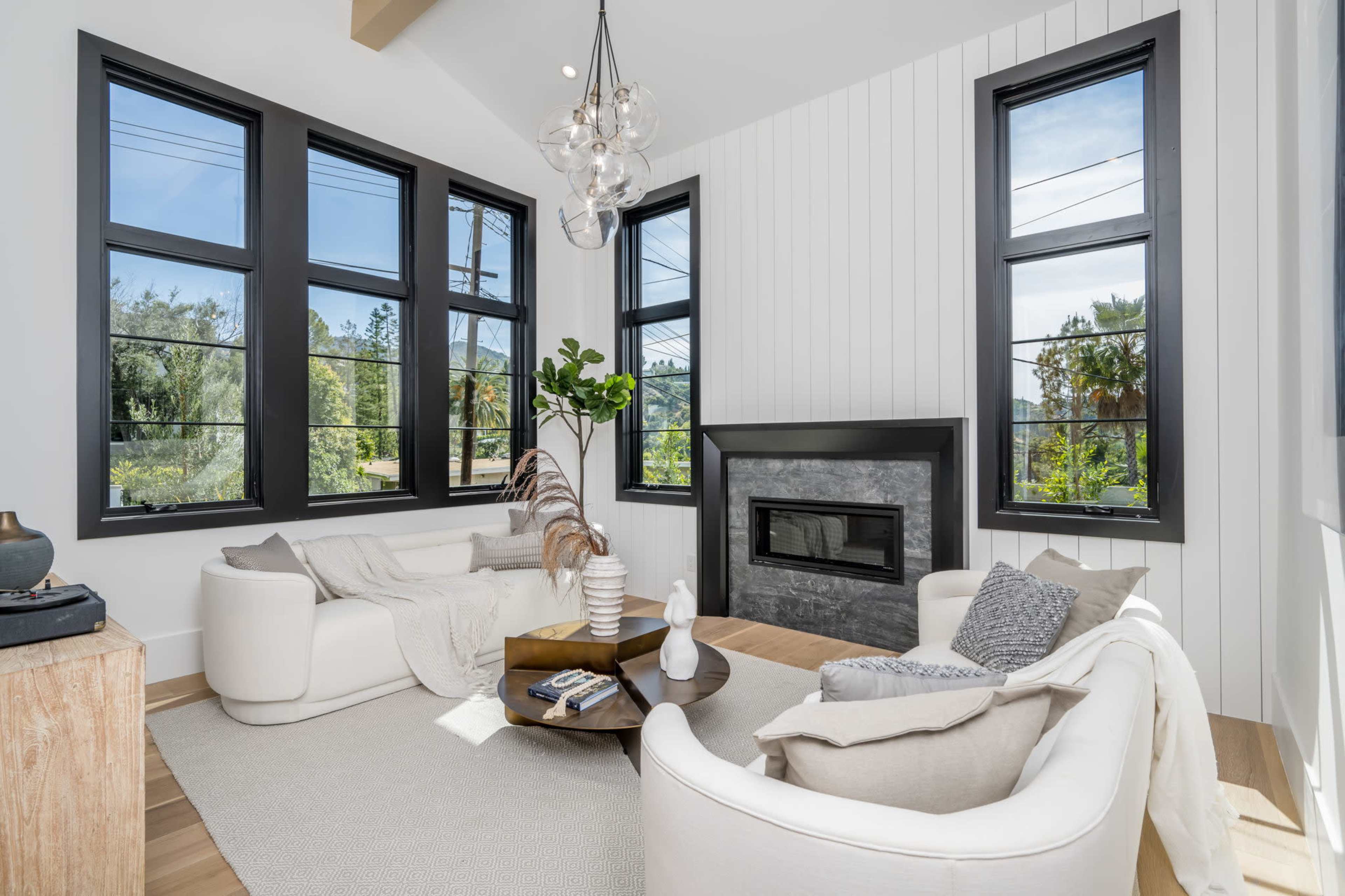 A modern living room features two white curved sofas, a round coffee table, and a fireplace, with large windows allowing natural light to fill the space.