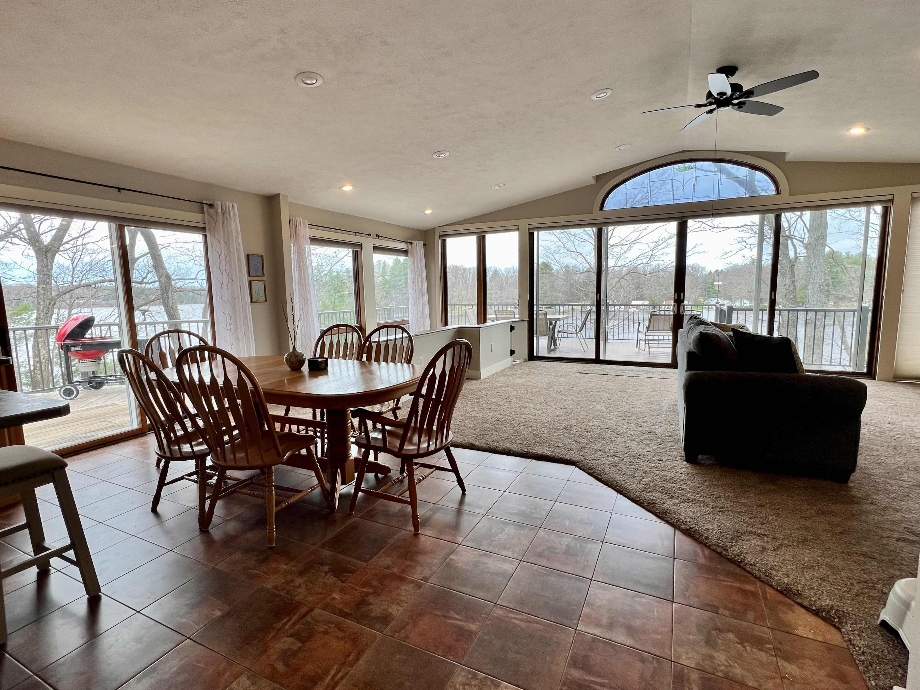 A spacious living area featuring a dining table with chairs, a couch, large windows overlooking a deck, and a view of trees outside.