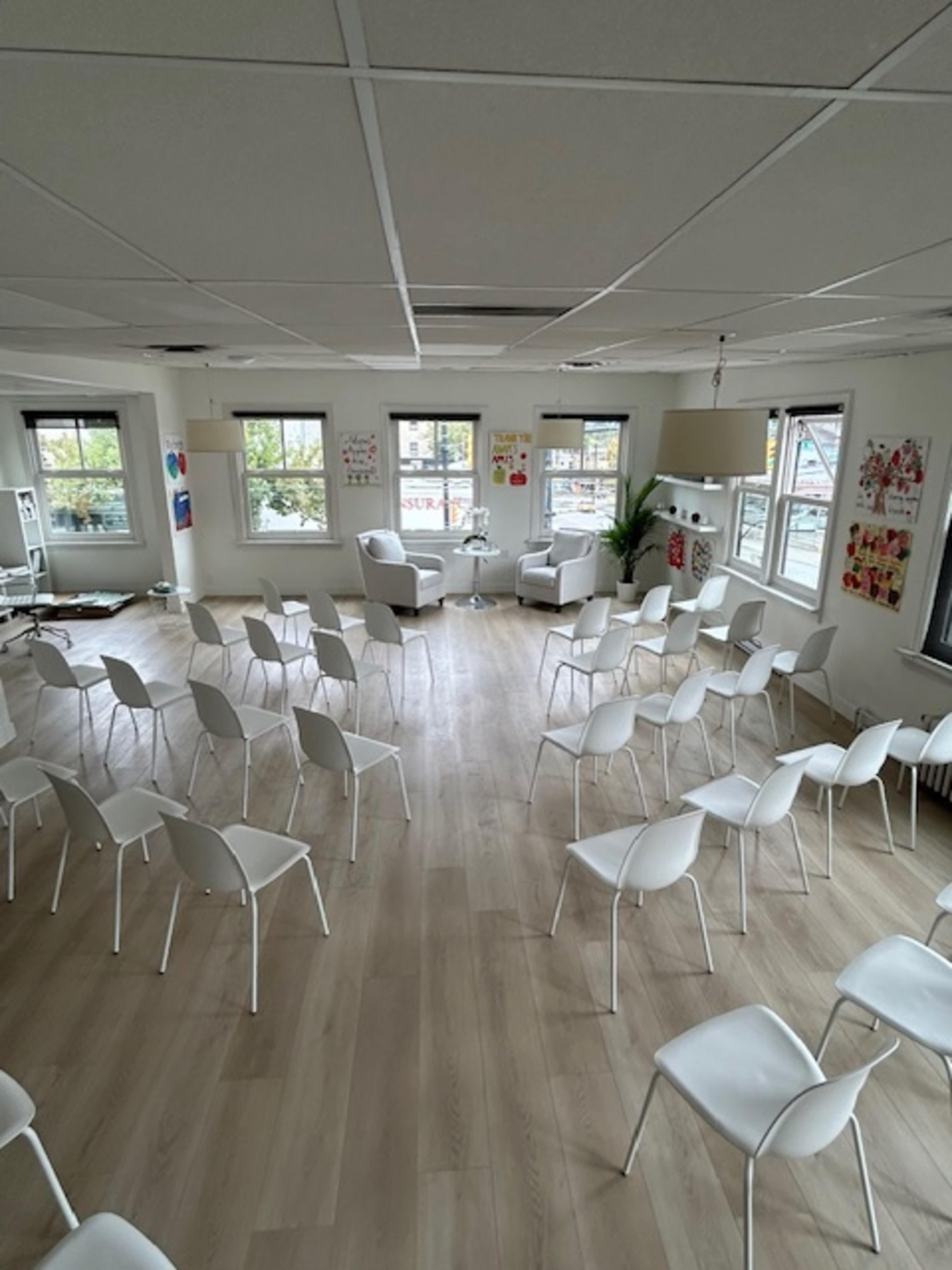 A spacious room with light wooden flooring features rows of white chairs arranged for an event, with large windows and colorful artwork on the walls.