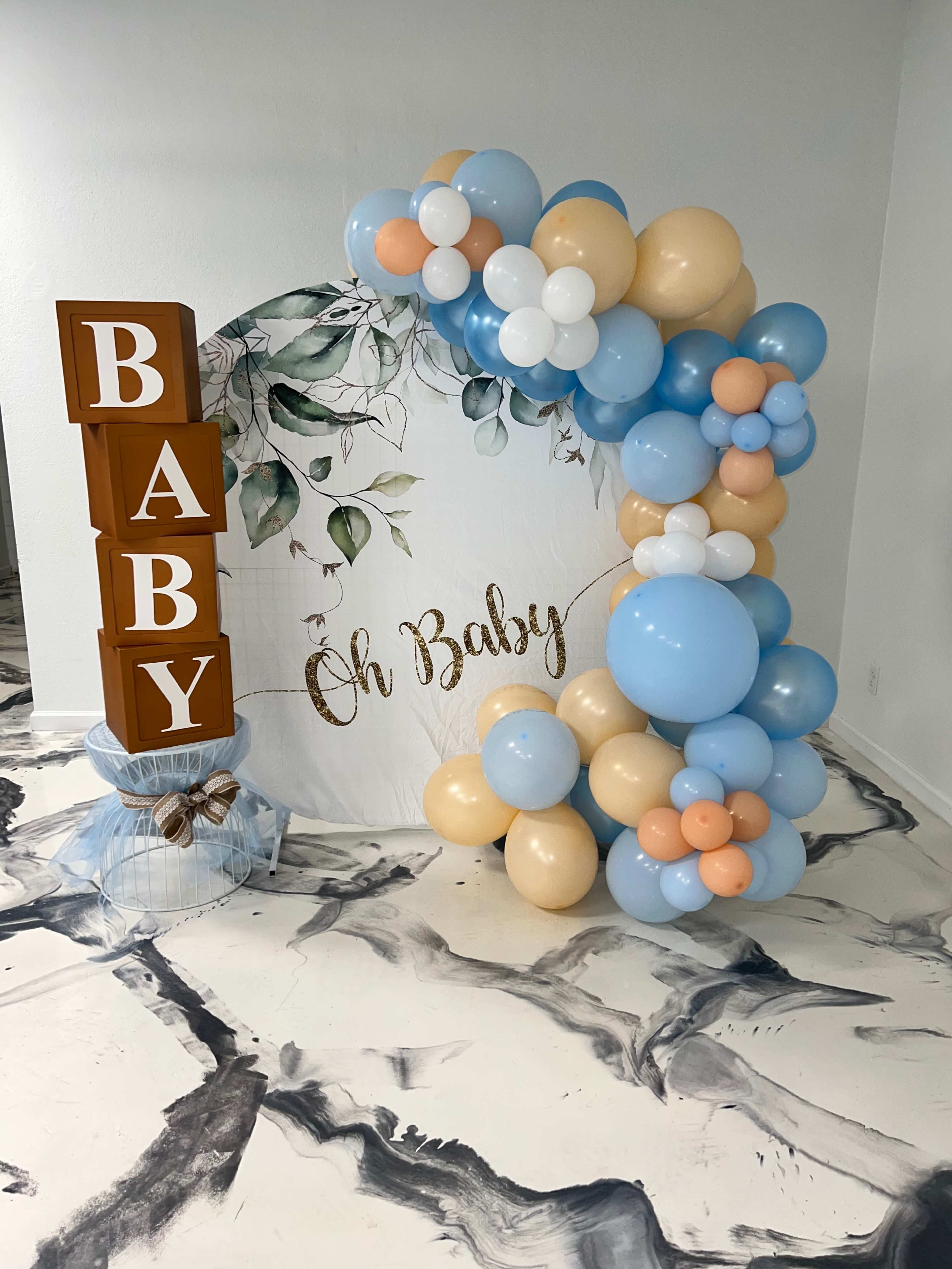 A decorative setup for a baby shower, including a large sign that reads "Oh Baby" and an arch made of blue, white, and beige balloons.