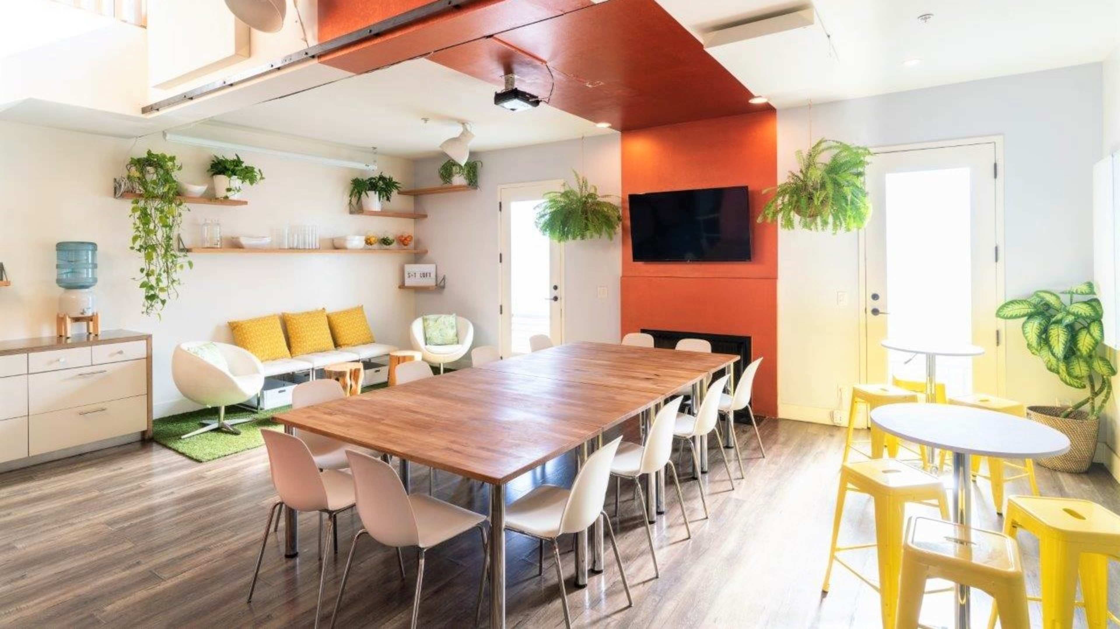 A modern communal space featuring a long wooden table surrounded by chairs, a cozy seating area with a sofa and plants, and bright yellow tables in a well-lit environment.
